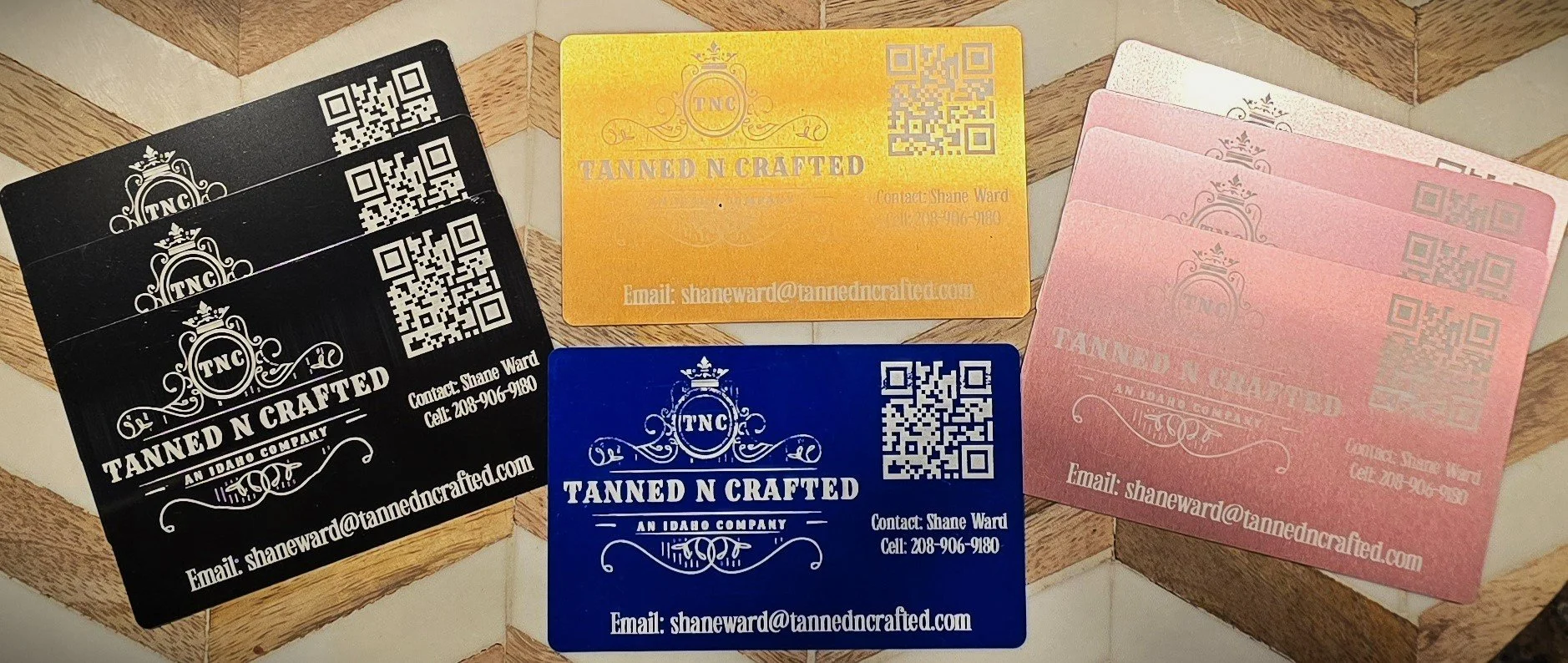 Stack of laser engraved business cards for Tanned N Crafted in black, yellow, blue, and rose gold colors, with a logo, QR code, contact information, and email address.