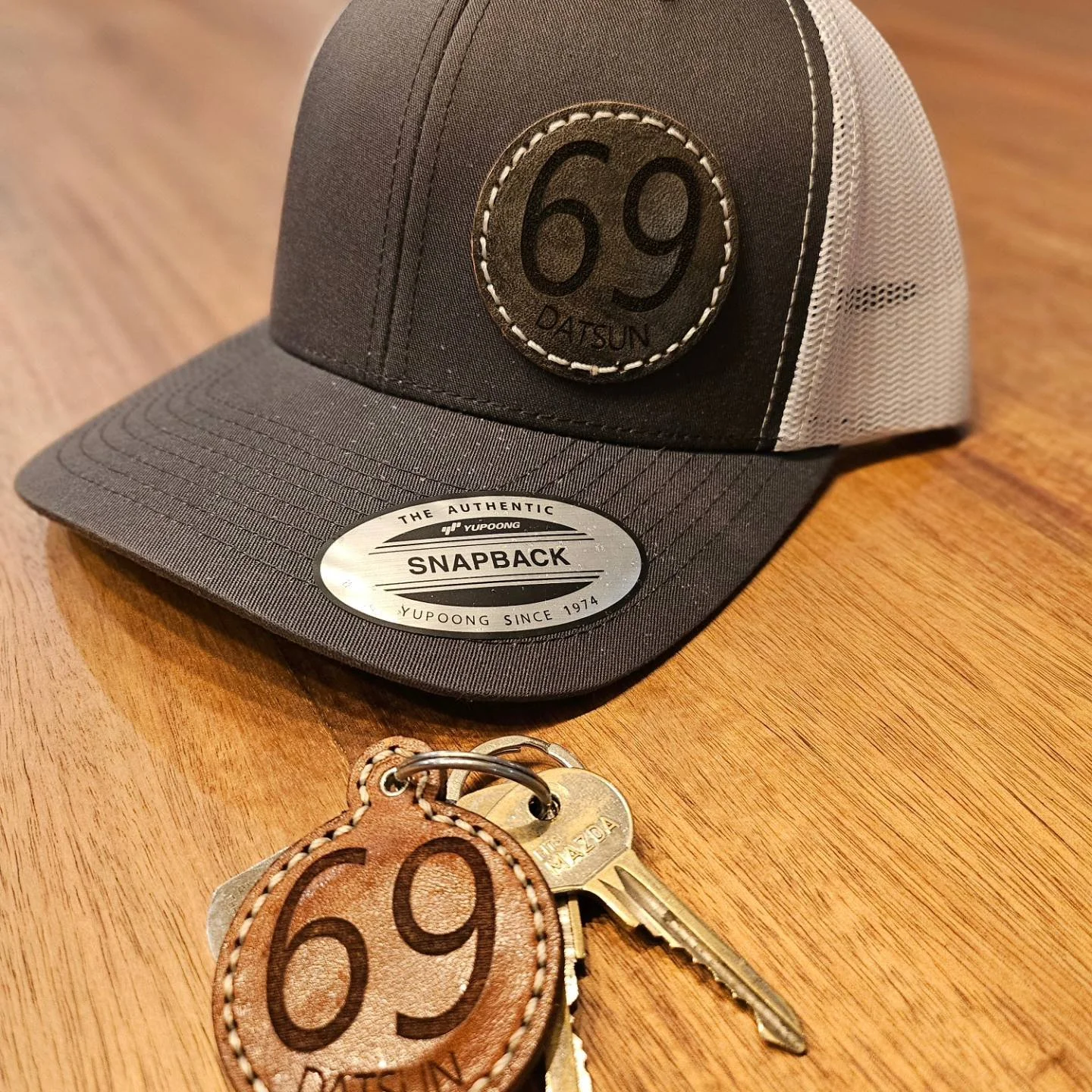 Gray and black trucker-style cap with custom laser engraved patch displaying the number 69 and the word DATSUN, and set of keys with a handsewn, laser engraved, matching leather keychain. 