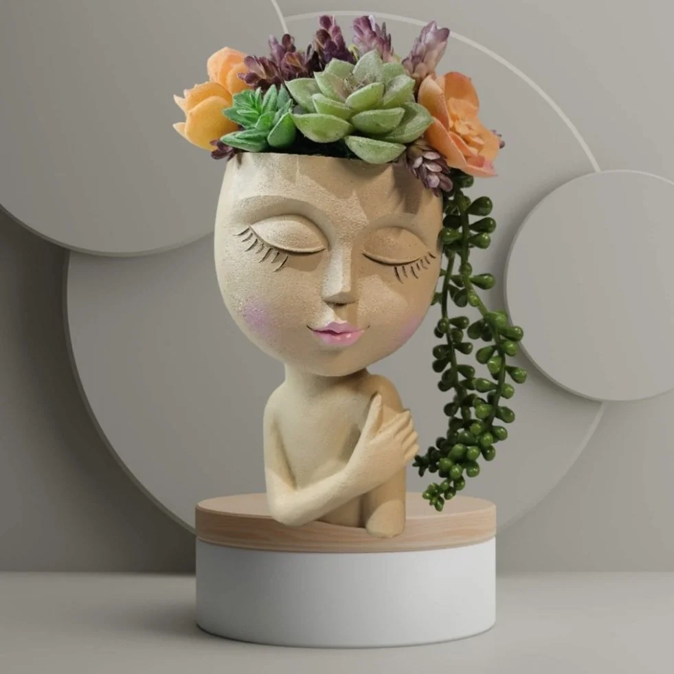 A decorative sculpture of a woman's face with closed eyes and pink lips, featuring a head made of various colorful artificial succulents and flowers, placed on a circular base on the counter in an office or business.
