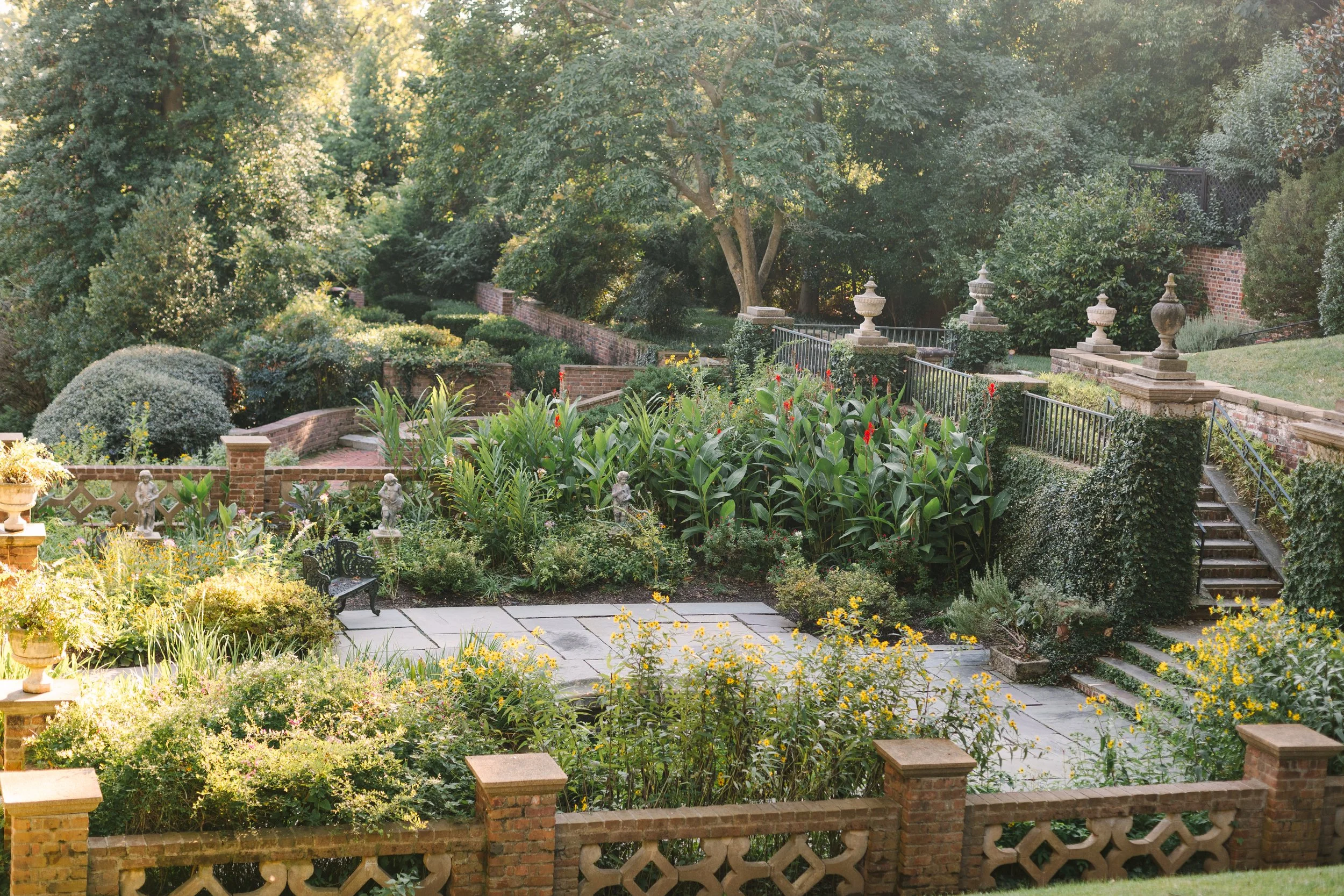 A lush, multi-tiered garden with brick walls, stairs, and decorative vases, filled with greenery, colorful flowers, and shrubbery, under bright sunlight.