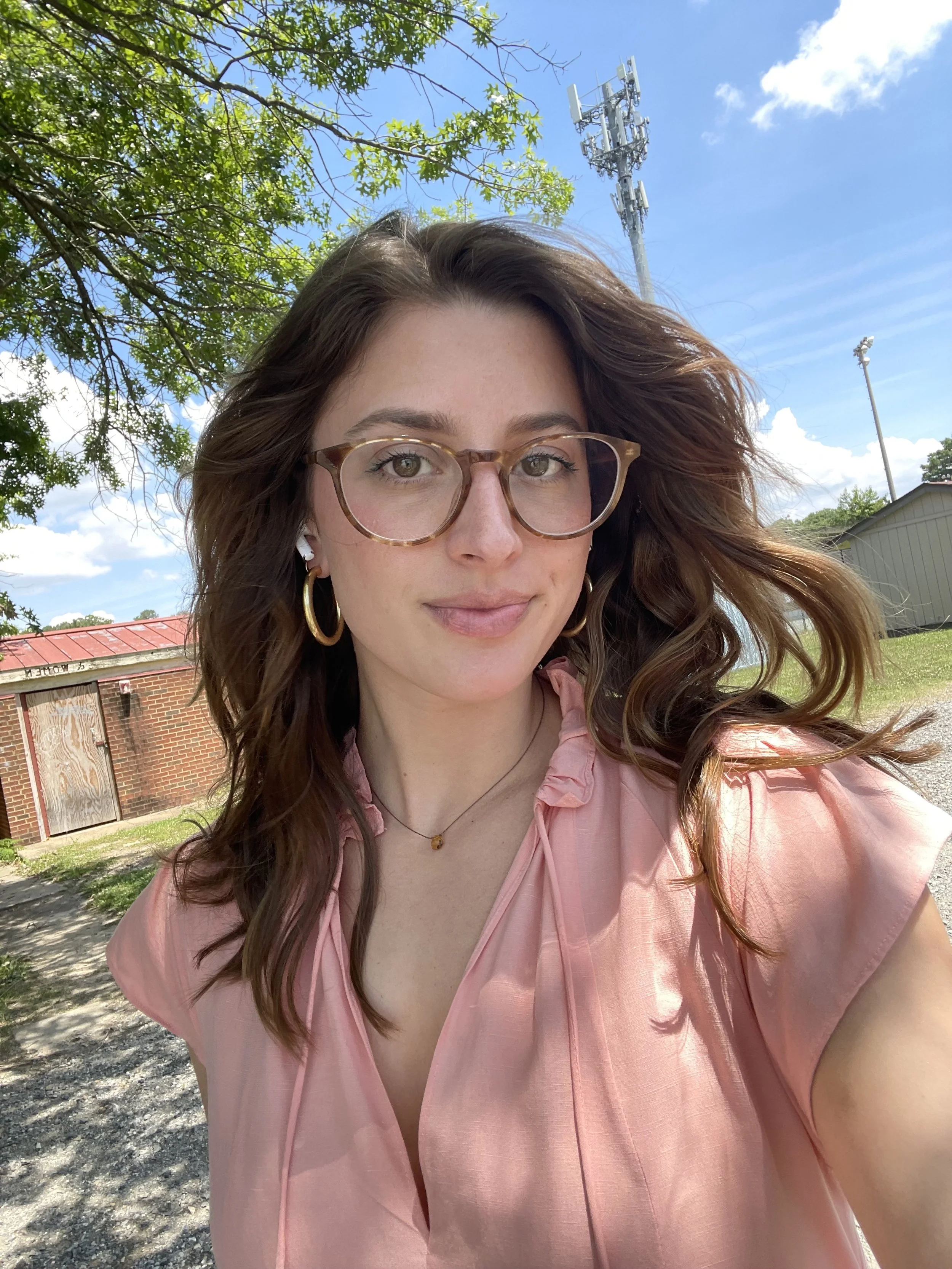 A young woman with wavy brown hair, glasses, and hoop earrings taking a selfie outdoors under a blue sky with some clouds, surrounded by green trees and a red brick building in the background.