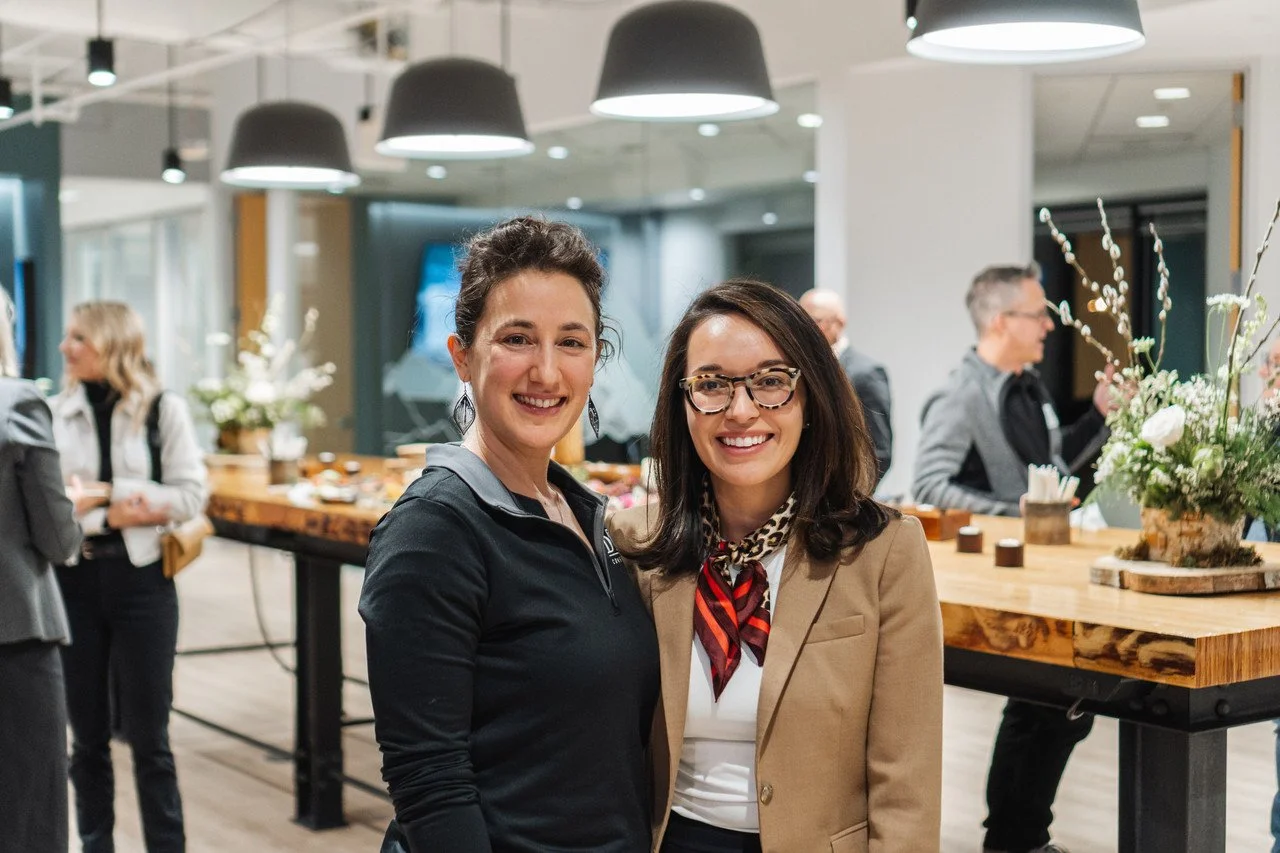 Two women smiling at a social event in a modern, well-lit room with a wooden table, floral arrangements, and pendant lights, with other people in the background.