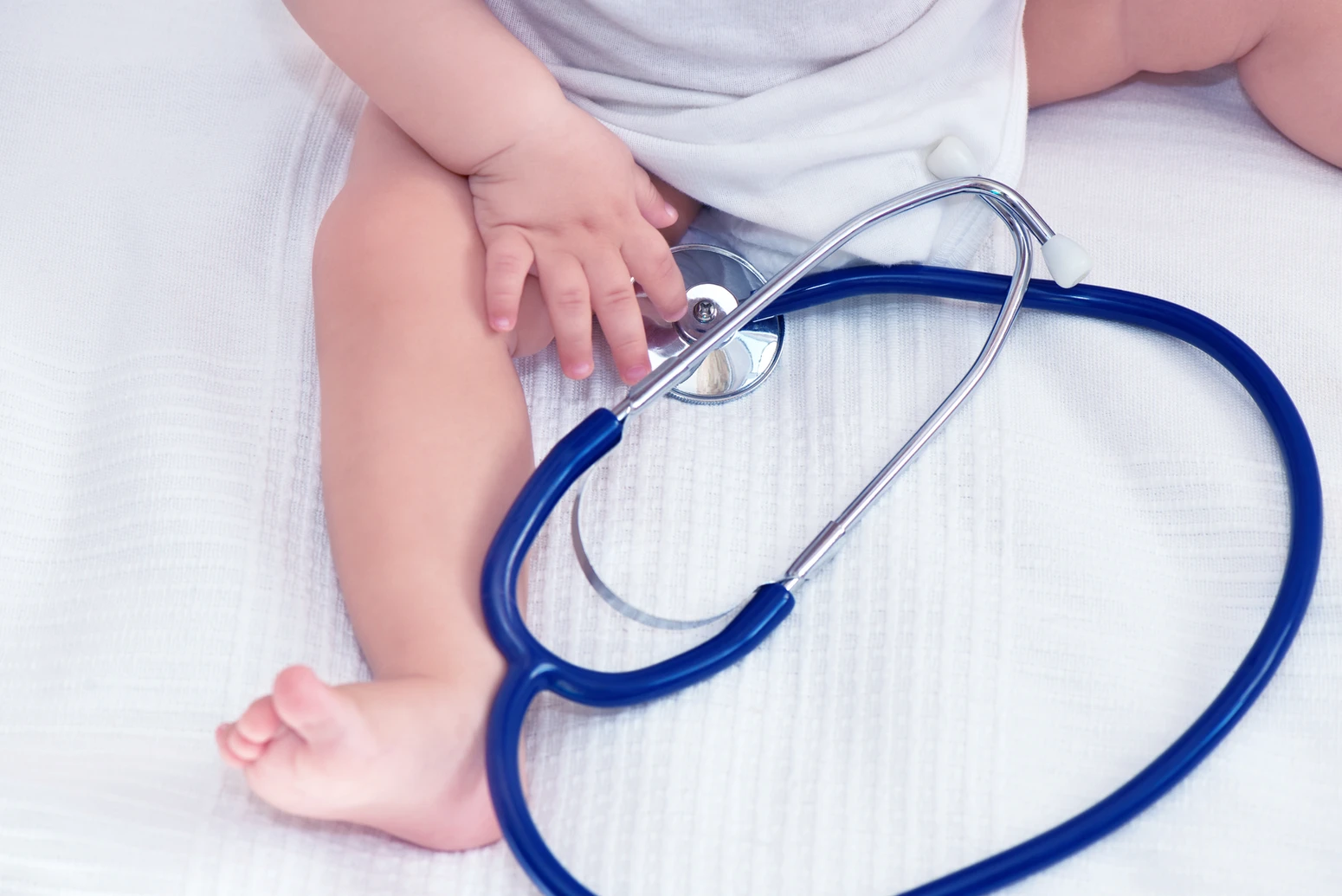 A baby sitting on a white blanket with their hands on a stethoscope