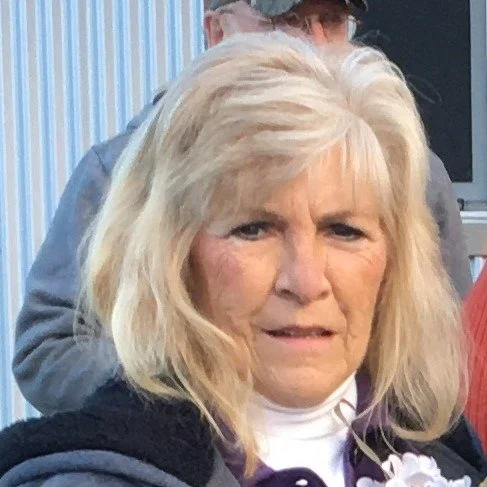 A portrait of an older woman with blonde hair, looking at the camera with a slight frown, in a setting with other people and vertical striped background.
