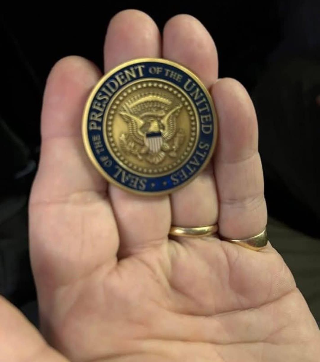 A hand holding a presidential seal badge of the United States with the text "The President of the United States" around the seal.
