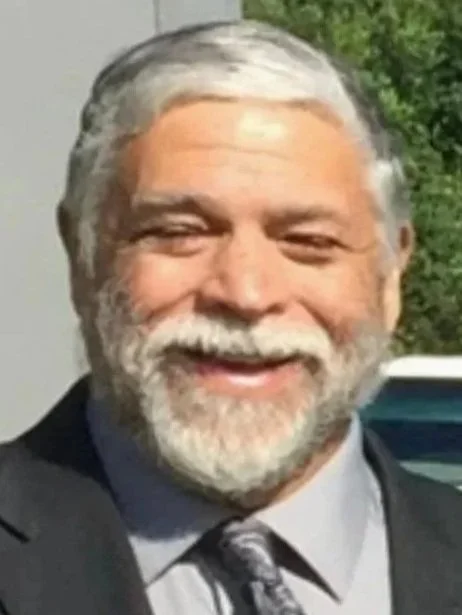 A smiling elderly man with gray hair and a beard, wearing a suit and tie, outdoors with greenery in the background.