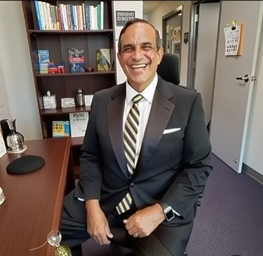 A man in a dark suit and striped tie sitting in an office chair, smiling at the camera, with a wooden bookshelf filled with books and decorative items behind him.
