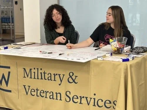 Two women sitting at a table with a yellow tablecloth that reads 'Military & Veteran Services.' one woman is writing and the other is reaching across the table.