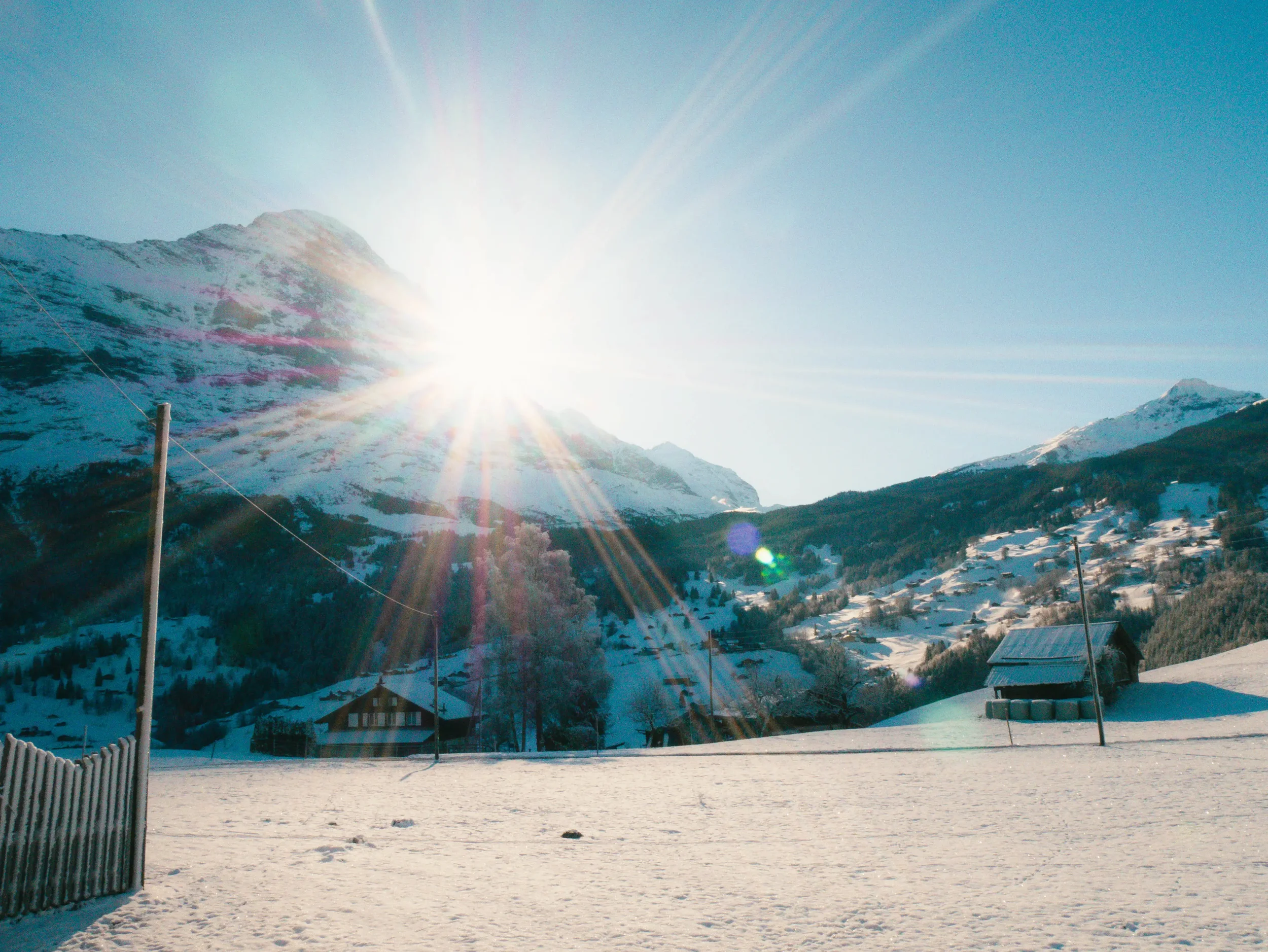 Schneebedeuchte Berge unter klarem Himmel mit der Sonne im Vordergrund, Sonnenstrahlen und lens flare, ländliche Häuser und Bäume im Tal.