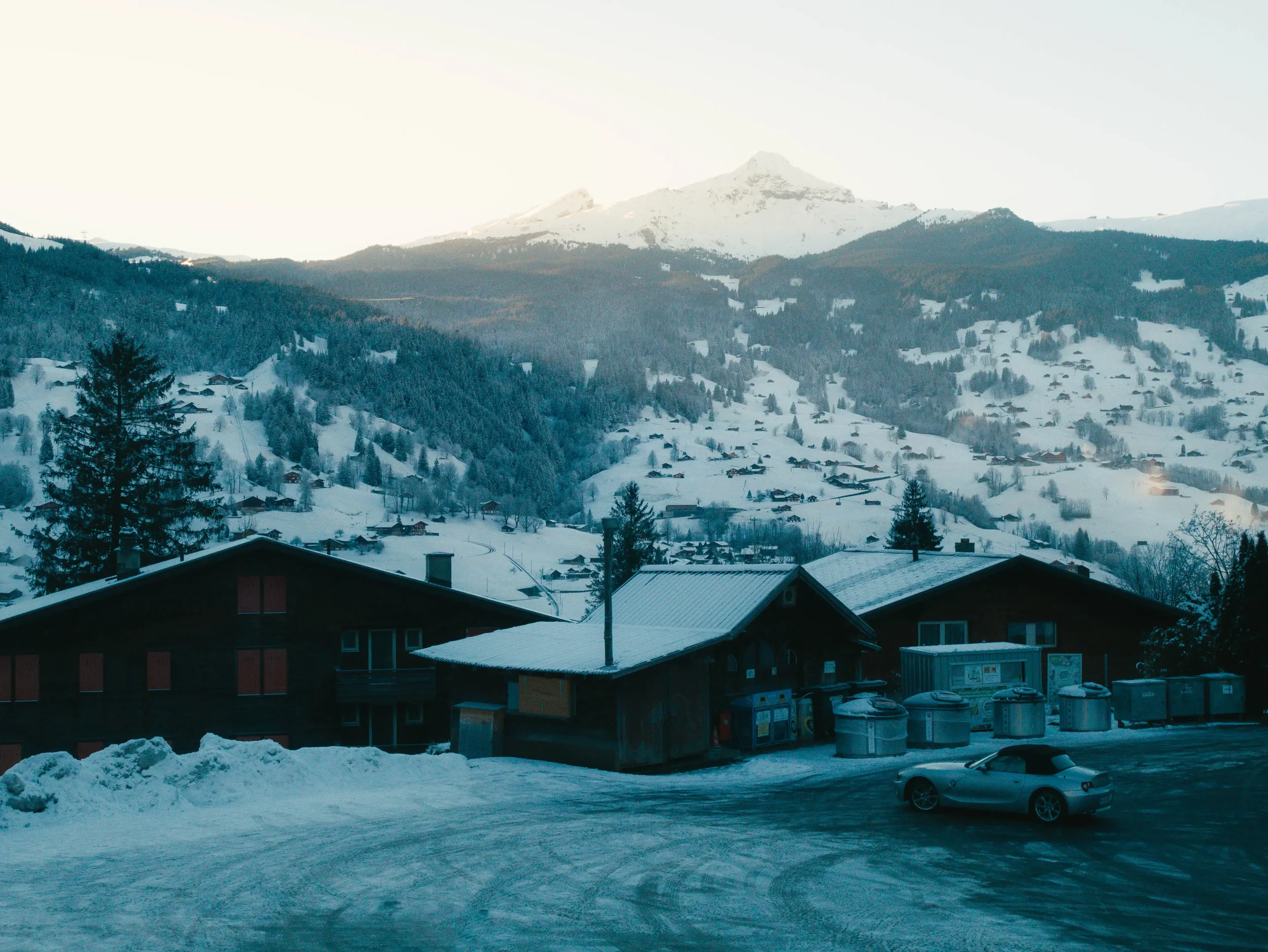 Schneebedeckte Landschaft mit Hütten, Bäumen und Bergen im Hintergrund, am frühen Morgen oder späten Nachmittag.