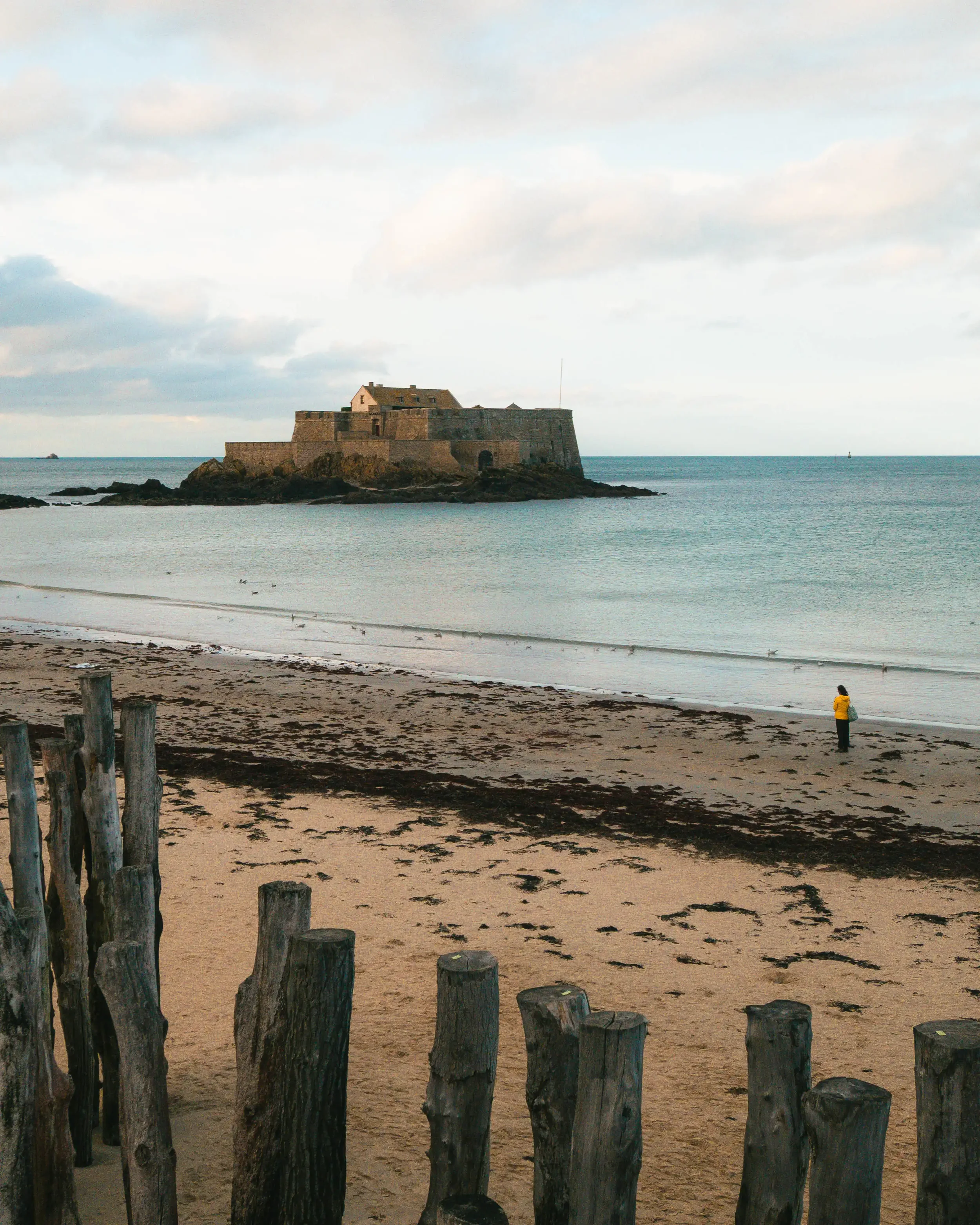 Ein einsamer Mensch steht am Strand, im Hintergrund ein alter Festung auf einer kleinen Insel im Meer, umgeben von Wasser und Himmel.