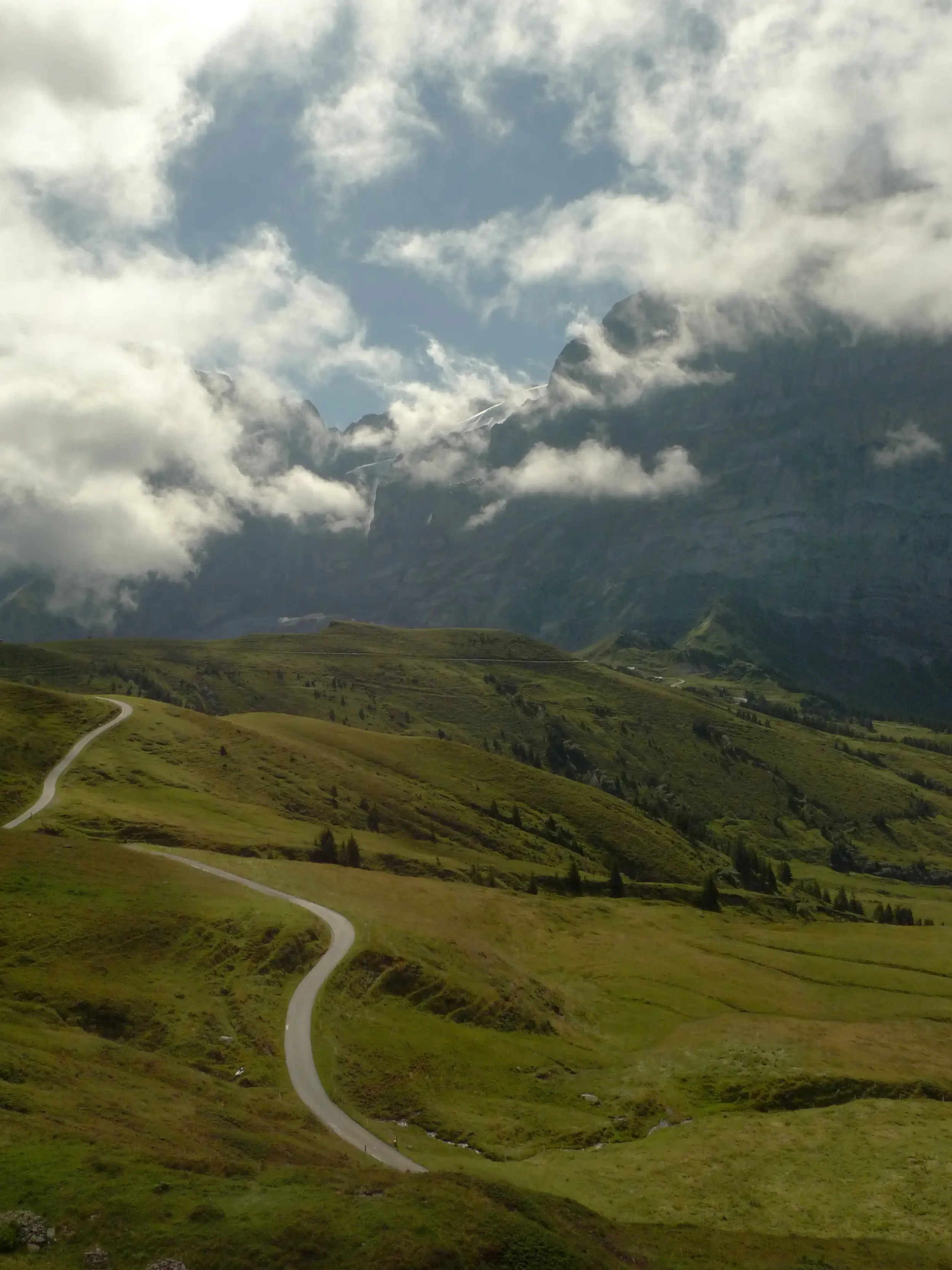 Grüne Hügel mit kurviger Straße, im Hintergrund hohe Berge mit Wolken und einer kleinen Schneeschicht auf einem Gipfel.