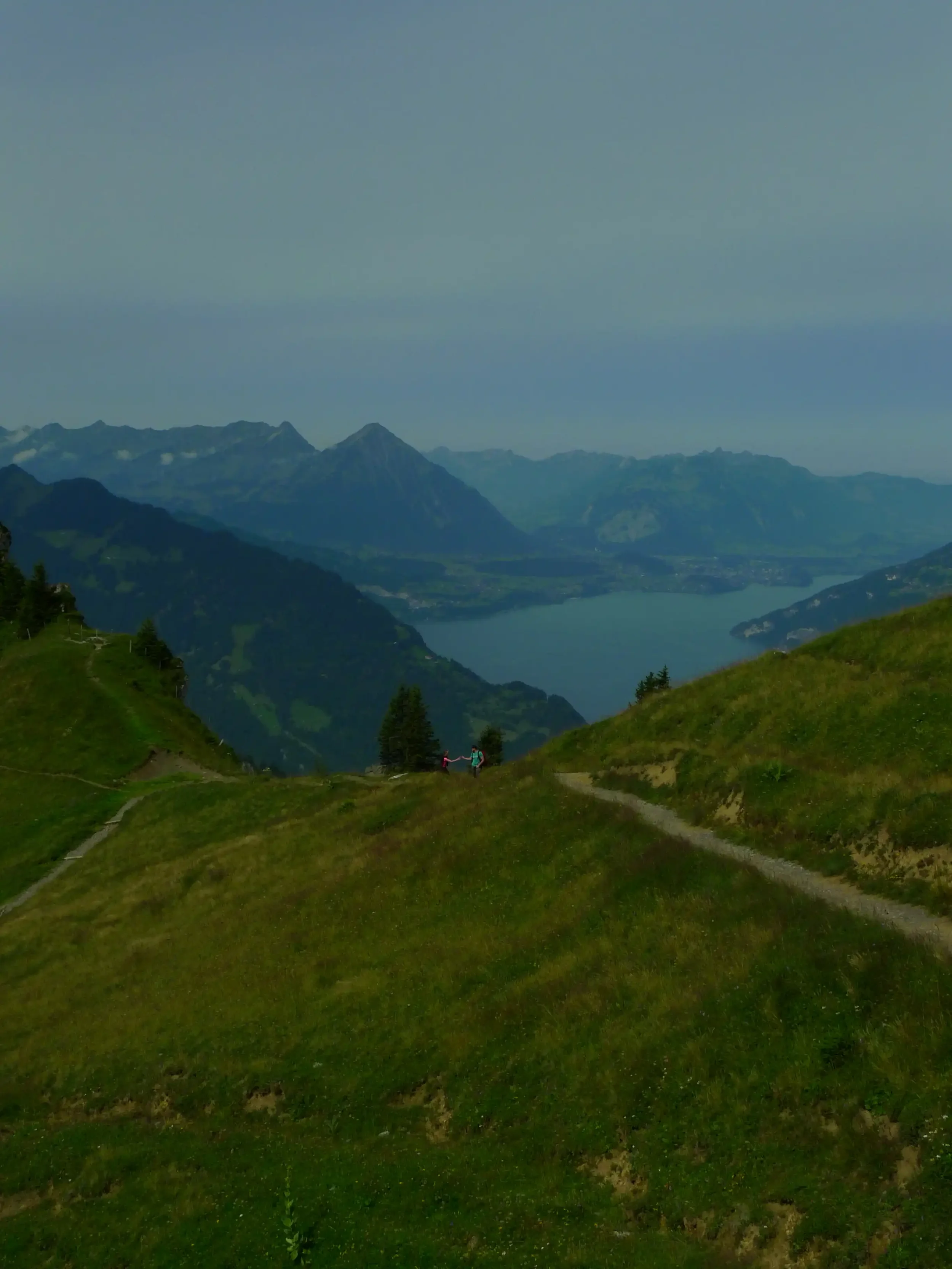 Zwei Personen auf einem Wanderweg in den grünen Hügeln mit Blick auf einen See und Berge im Hintergrund.