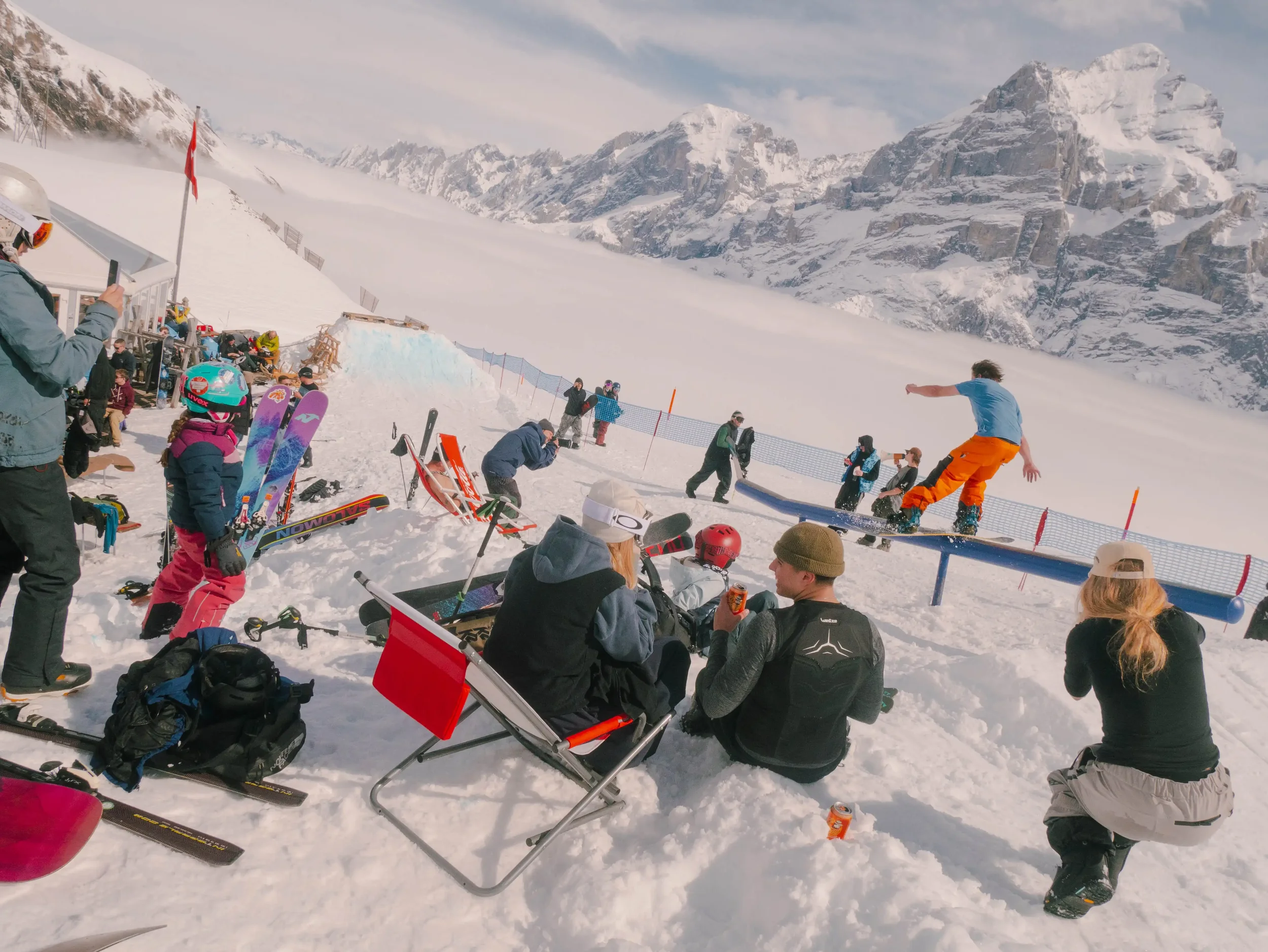 Ski- oder Snowboardfahrer zeigt einen Trick auf einer Schneehalbpipe, umgeben von Skifahrern und Zuschauern im Winterskigebiet mit Schneebergen im Hintergrund.