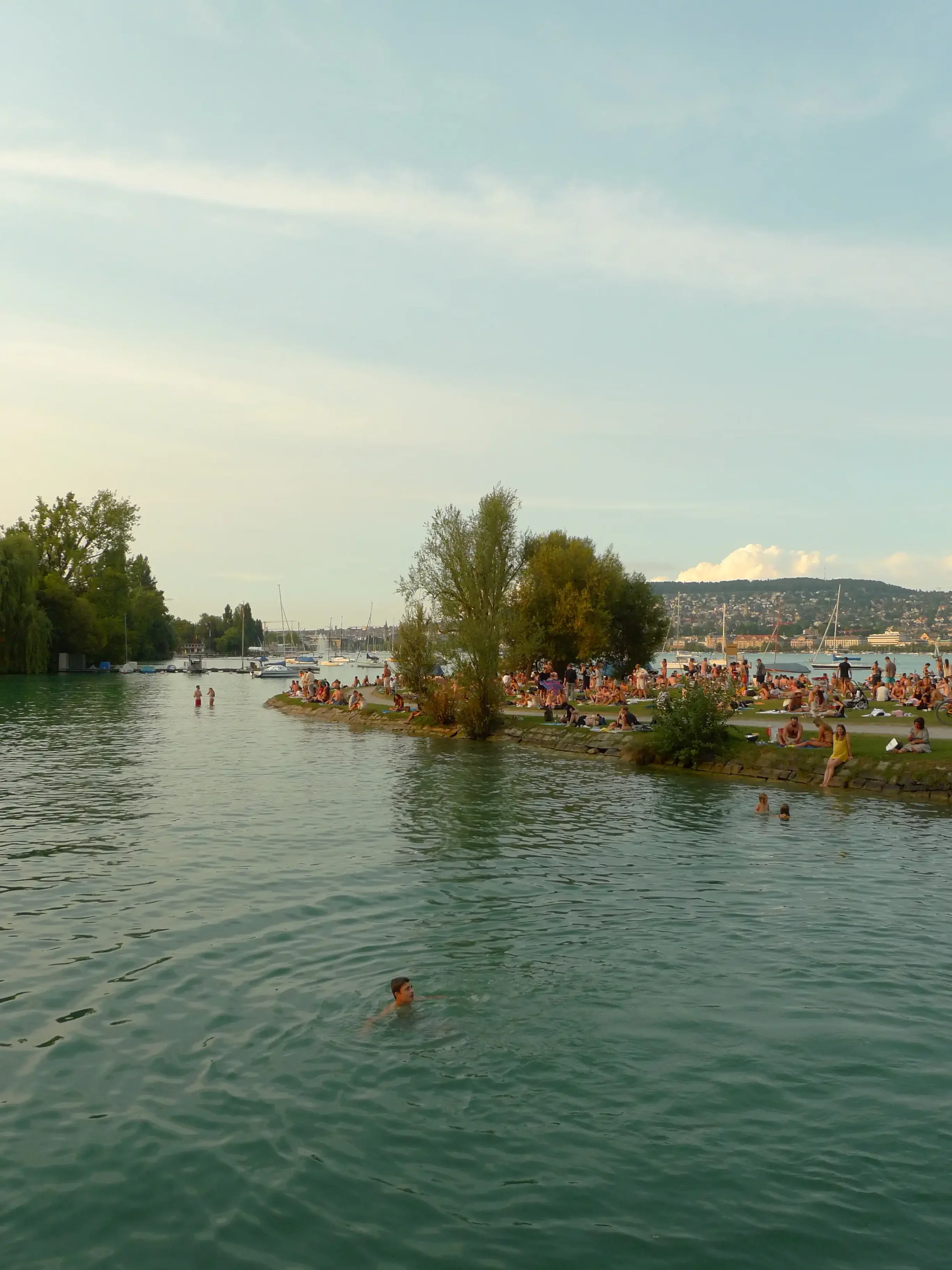 Menschen entspannen am Ufer eines Fluss- oder Seelands, einige schwimmen im Wasser, andere sitzen auf dem Gras, im Hintergrund befinden sich Boote und ein Stadtbild mit Hügeln.