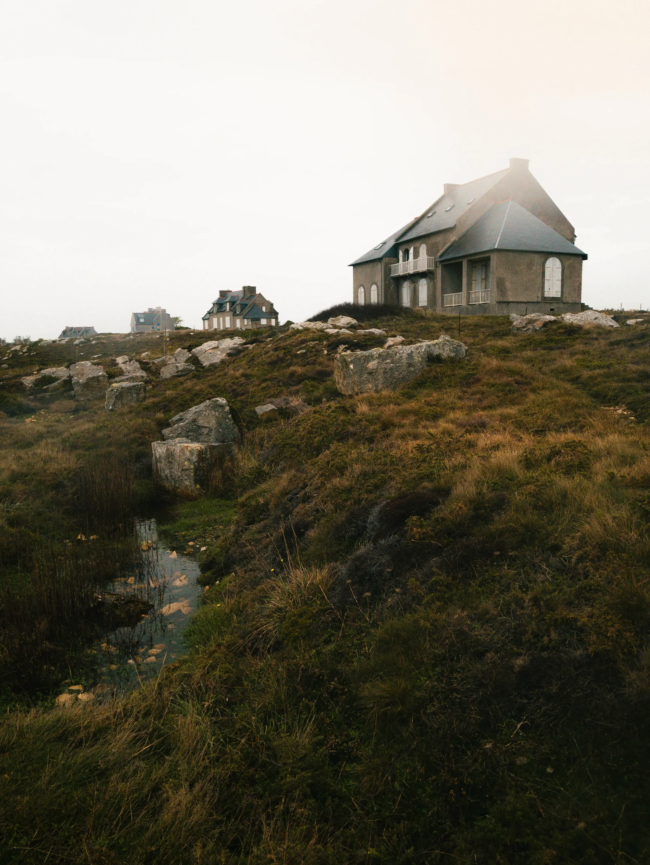 Ein abgelegenes Haus auf einem Hügel mit mehreren Fenstern und einem Balkon, umgeben von einer grasbewachsenen, felsigen Landschaft mit einem kleinen Wasserlauf im Vordergrund.