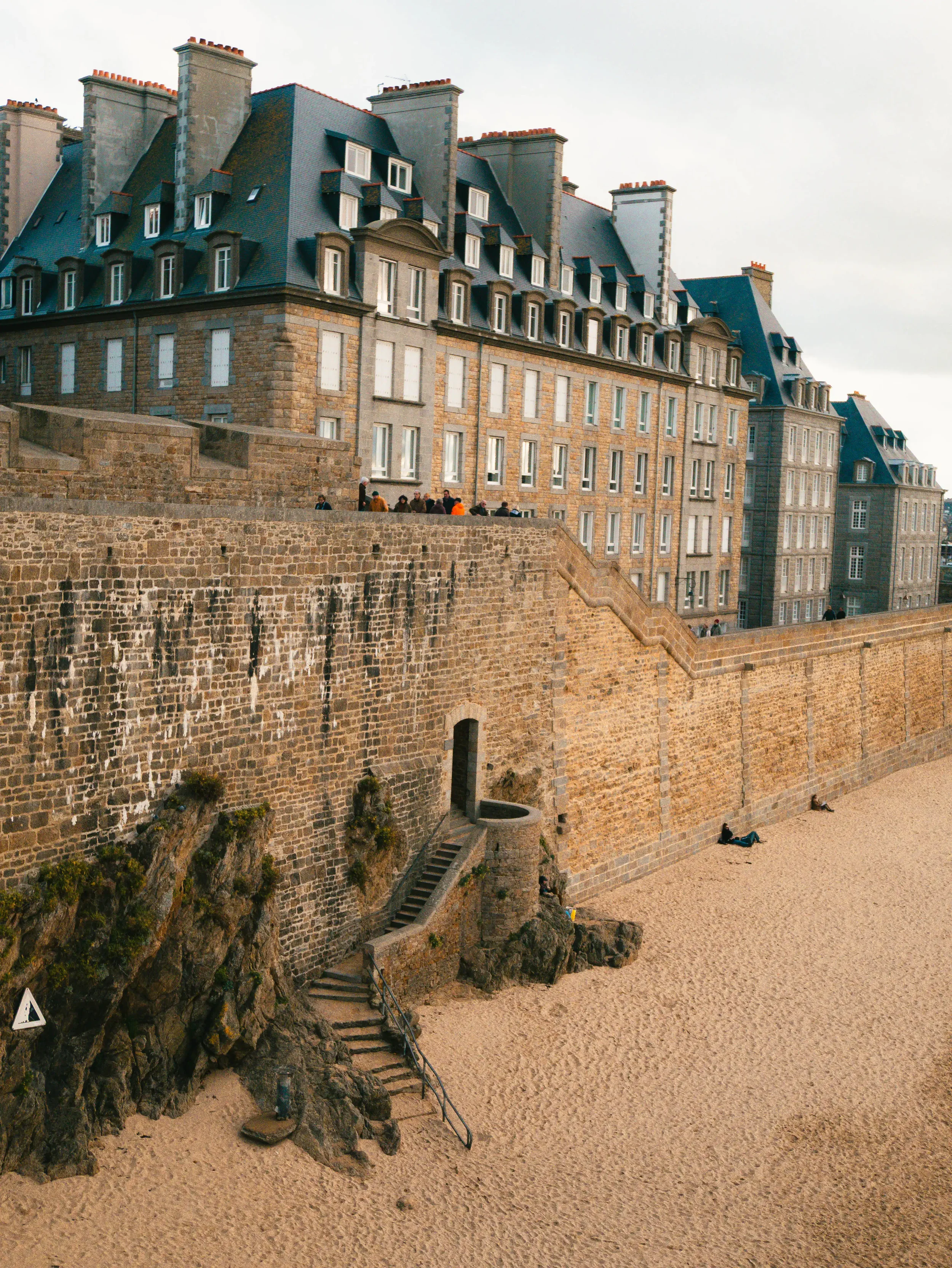 Historisches Gebäude an einer Küste mit Sandstrand, ein Steg führt von der Straße auf den Sand, Menschen sitzen und liegen am Strand.