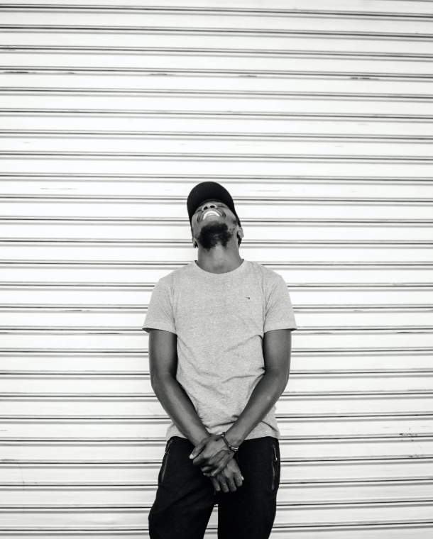 Young man standing in front of a closed metal shutter, smiling and looking up, wearing a cap, t-shirt, and pants.