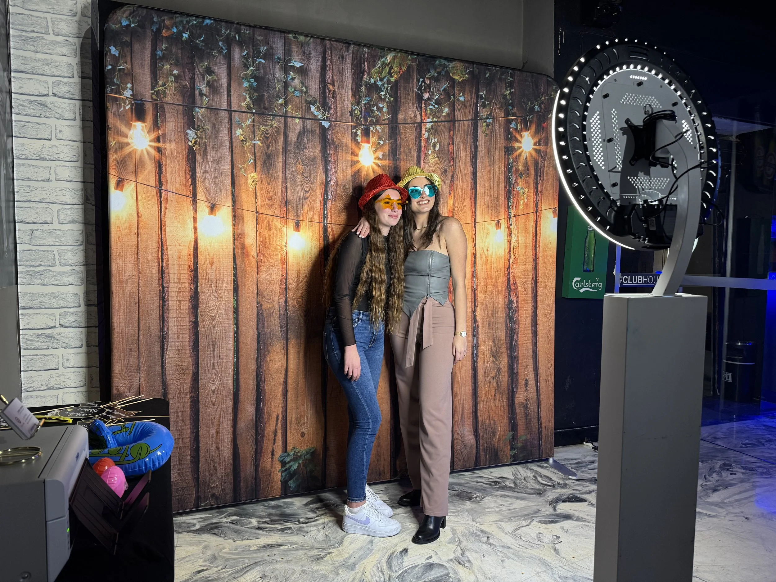 Two people posing with colorful glasses in front of a wooden backdrop with string lights in a photo booth setup.