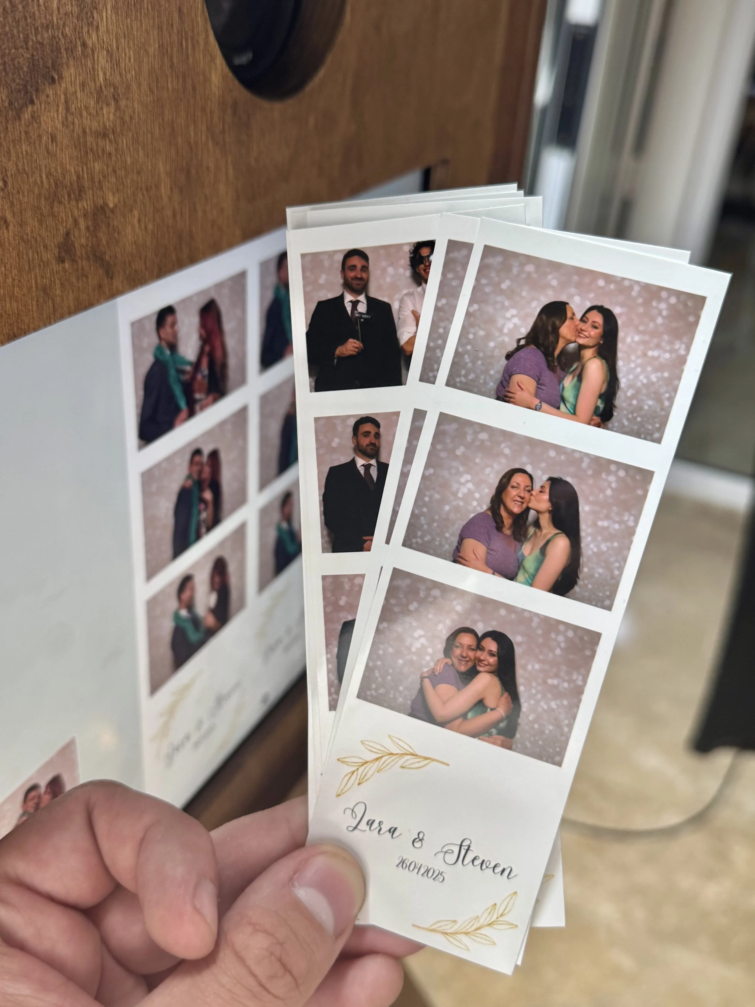 Hand holding photo booth strips of smiling people at an event, with names Lara & Steven dated 26042025 printed on them.