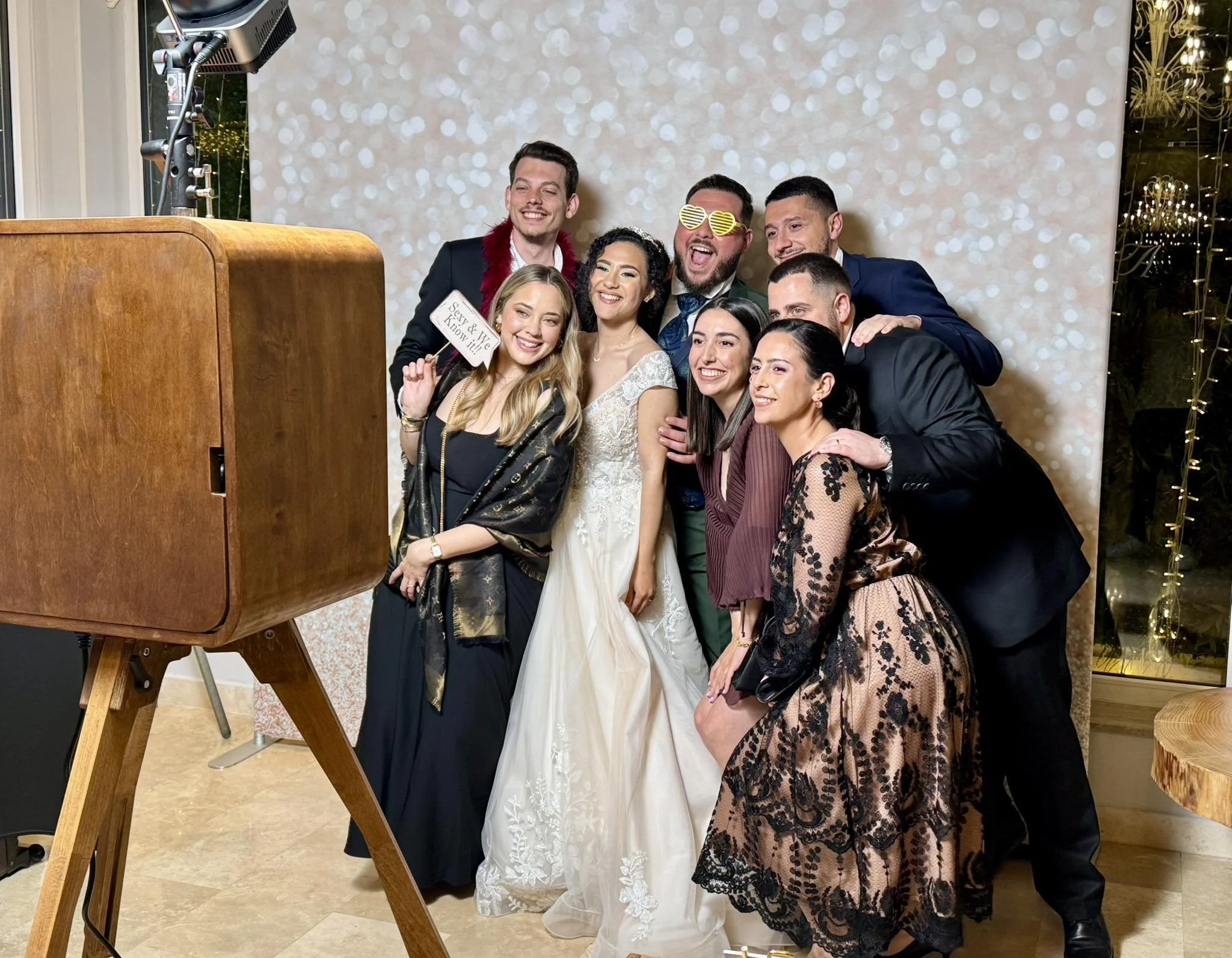 Group of people posing for a photo booth at a wedding, including a bride in a white gown, with a decorative background and a vintage wooden camera setup.