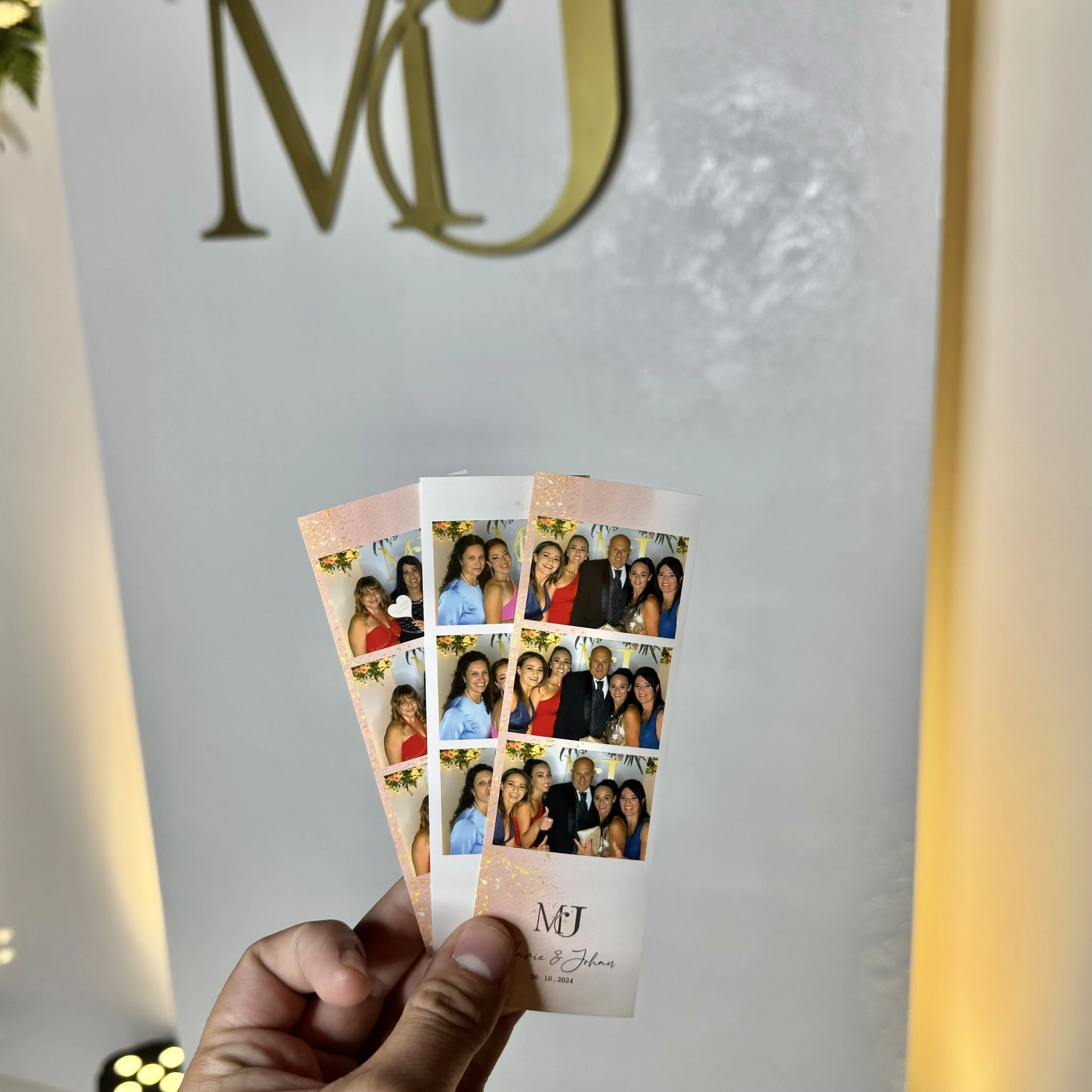Person holding two photo strips from a photo booth, featuring multiple people smiling and posing together. The background has gold letters 'MJ' and floral decorations, suggesting a celebration or event.