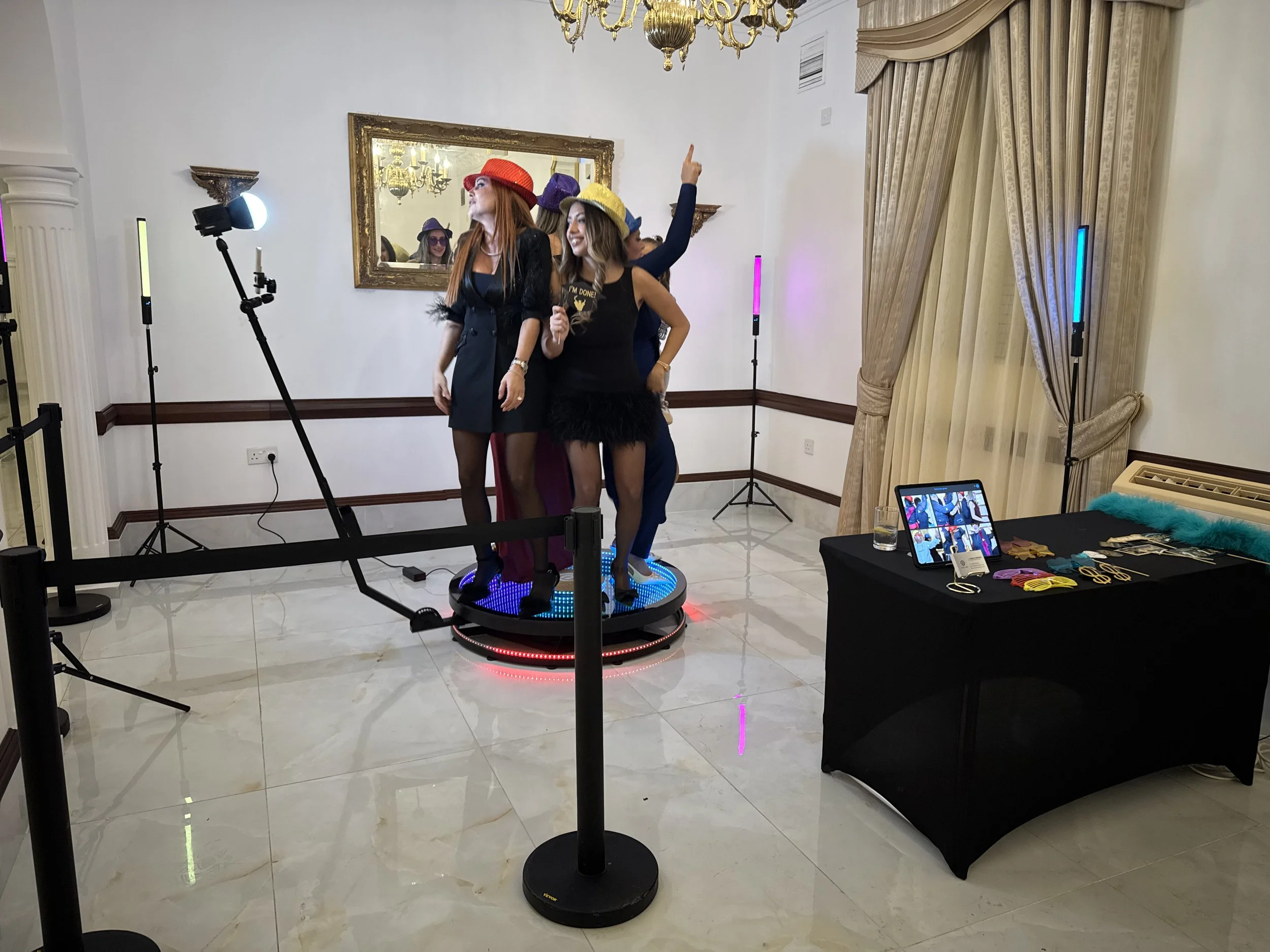 People posing on a 360-degree photo booth platform with lights and accessories in a decorated room.