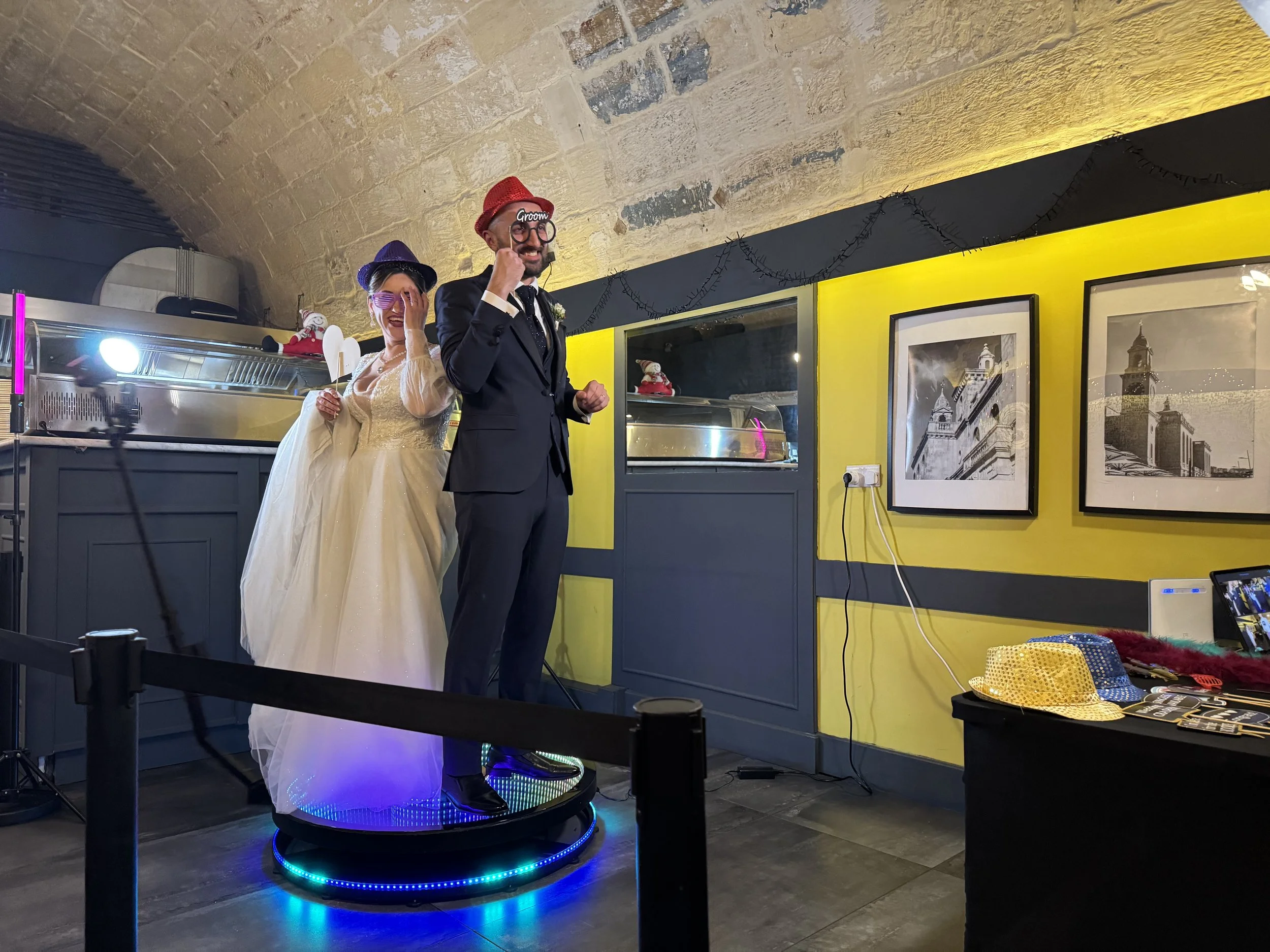 A bride and groom stand on a rotating platform in a decorated room. They are wearing colorful hats and playful glasses, posed with fun props. The bride is in a white dress, and the groom is in a dark suit. The background includes framed pictures on a yellow wall and various event decorations.