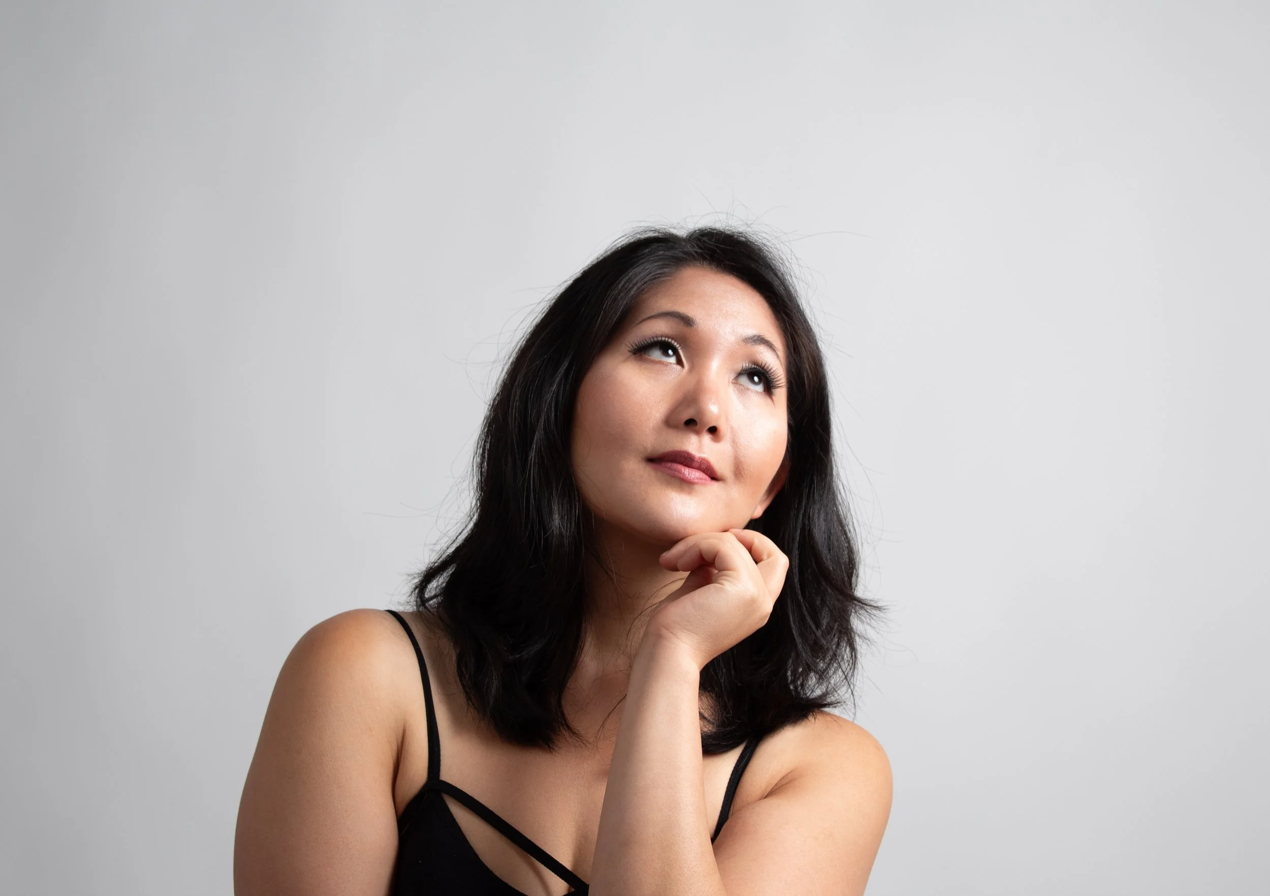 A woman with dark hair and a black top looking upward thoughtfully, resting her chin on her hand in front of a plain gray background.