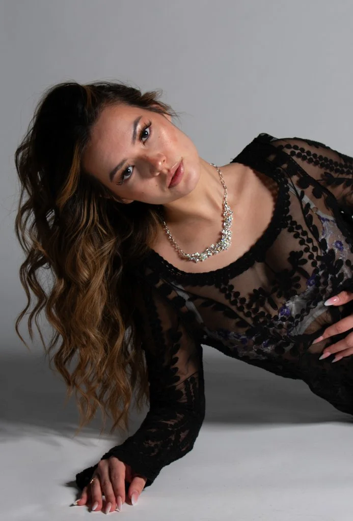 A woman with long wavy hair lying on the floor, wearing a black lace top and a silver necklace, looking at the camera.