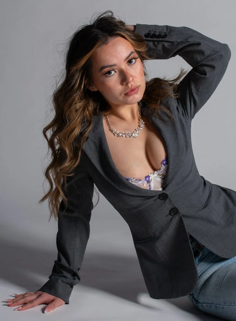 A woman with long wavy hair wearing a gray blazer, purple and white lace top, and a silver necklace poses against a neutral background