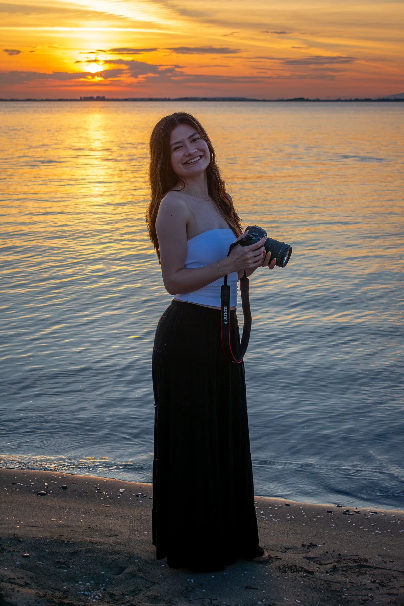 A woman with long hair smiling and holding a camera on a sandy beach during sunset with orange and yellow sky and calm water in the background.
