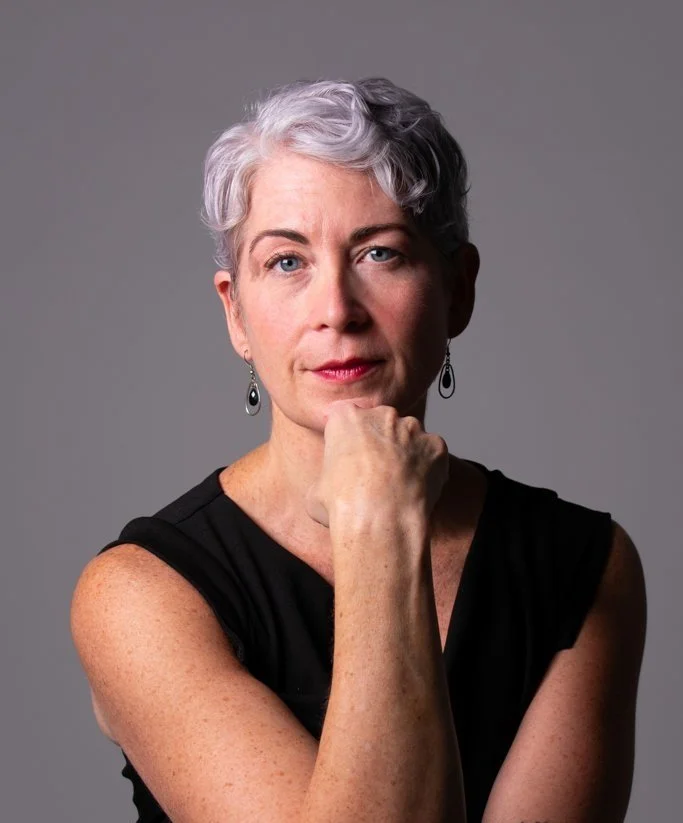Portrait of a mature woman with short gray hair, wearing a black sleeveless top and earrings, resting her chin on her hand against a neutral gray background.