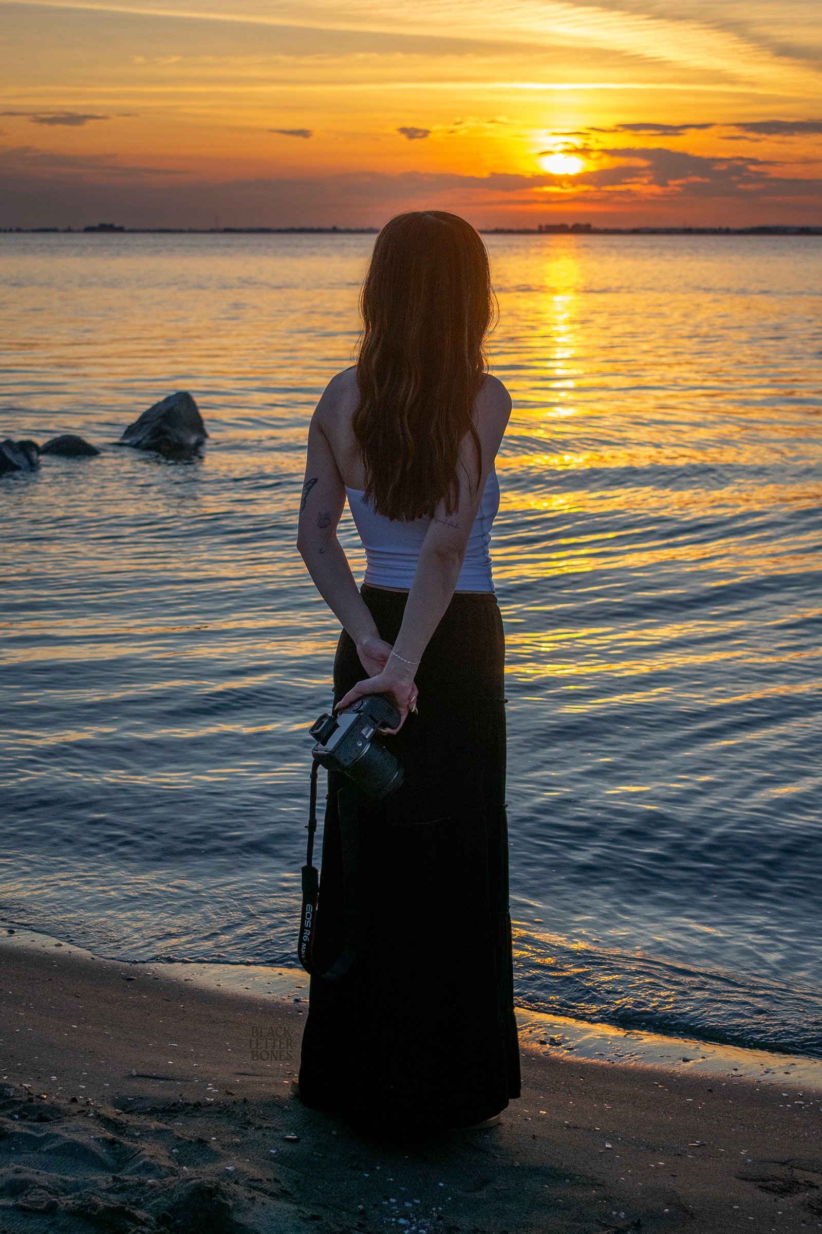 A woman standing on the beach at sunset, holding a camera behind her back, facing the water, with a colorful sky and calm waves.