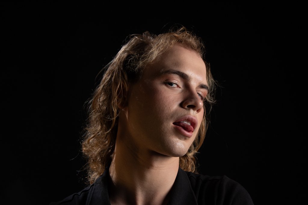 A young man with long, wavy blonde hair against a black background, looking at the camera with his tongue slightly out.