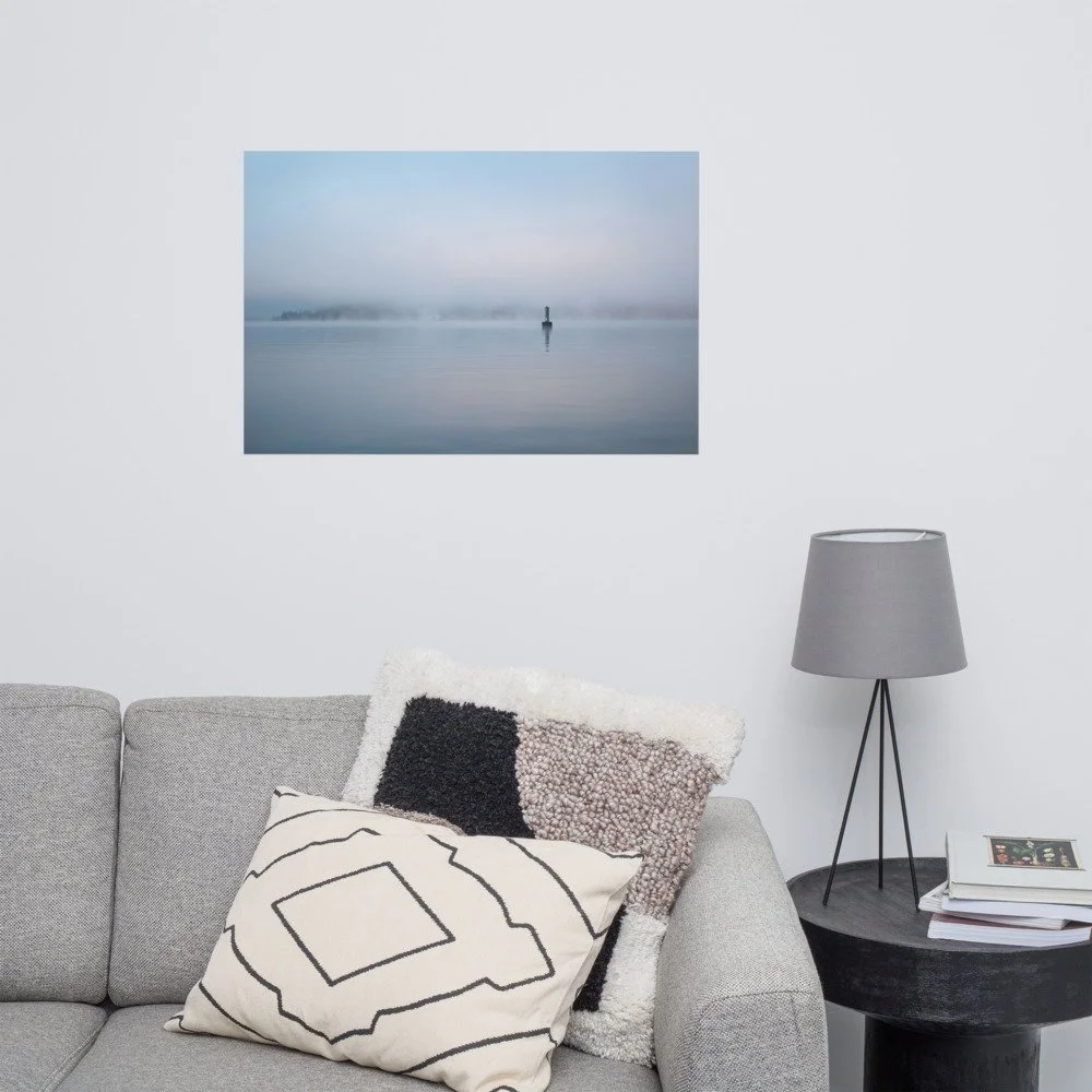 Living room with grey sofa, textured throw pillows, a side table with books, and a grey lamp. A framed photograph of a foggy lake scene hangs on the white wall.