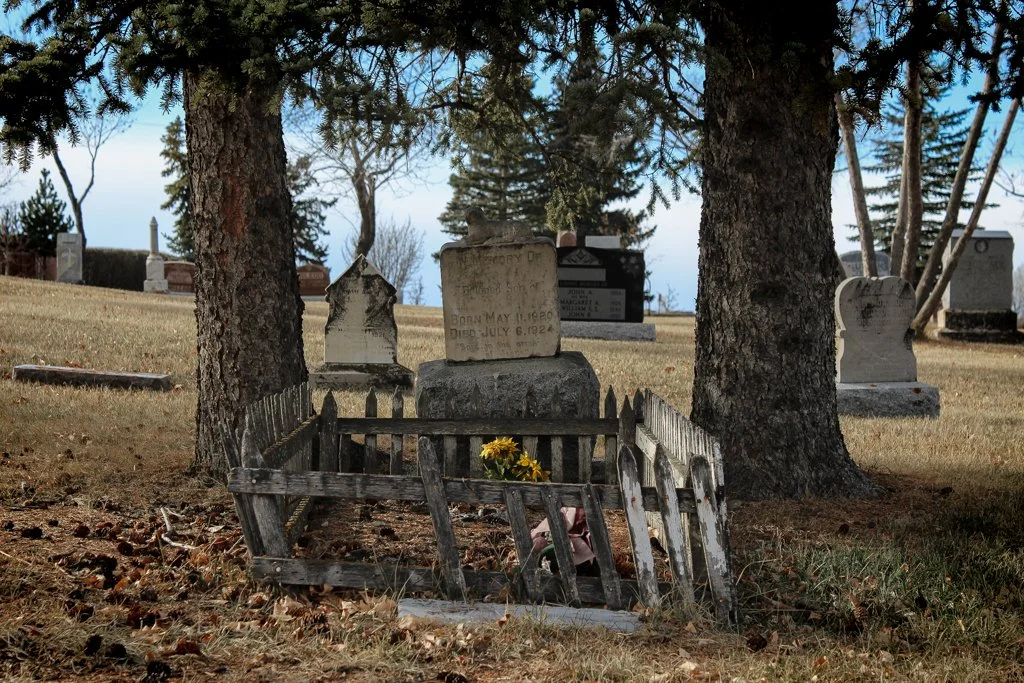 A broken wooden fence surrounds a small grave with yellow flowers in a cemetery with mature trees and various tombstones in the background.