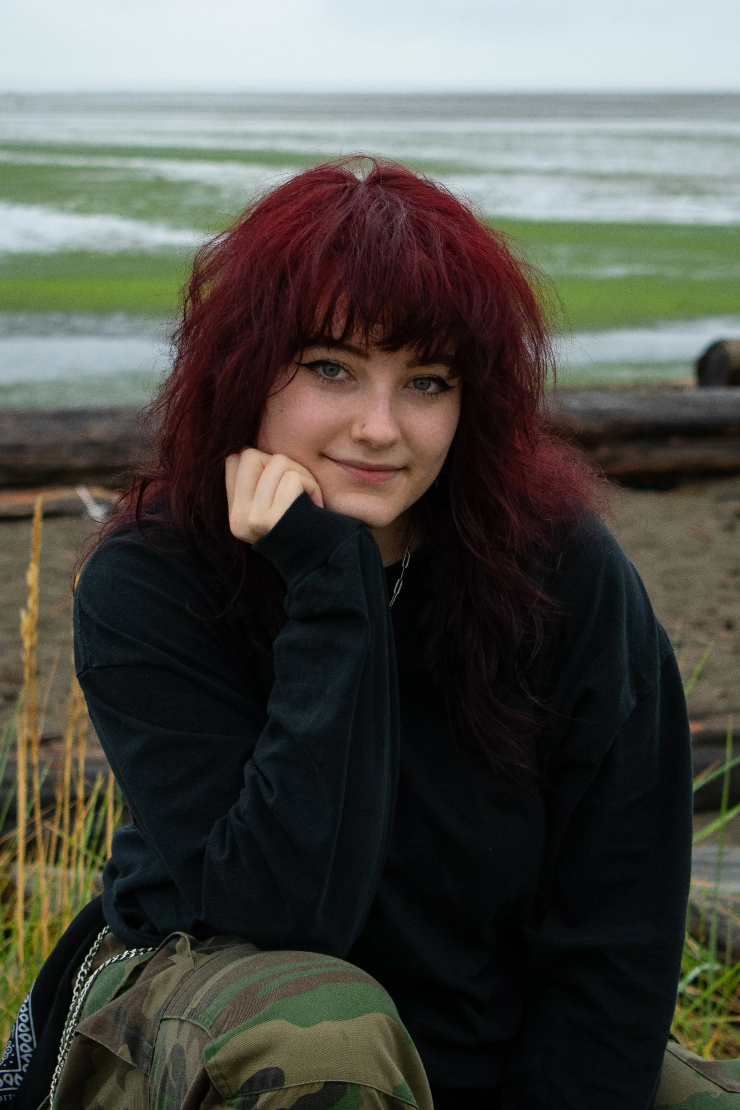A young woman with red hair and blue eyes sitting on a beach with grass and driftwood, wearing a black hoodie and camouflage pants.