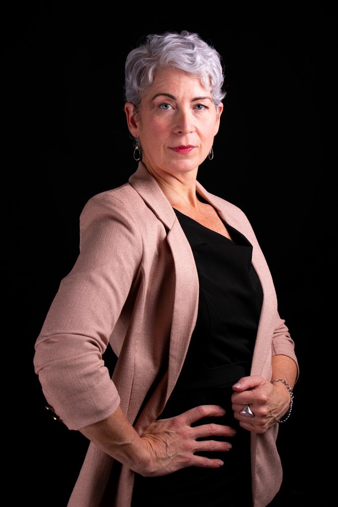 An older woman with short, curly silver hair wearing a light pink blazer and black top, standing confidently against a black background.