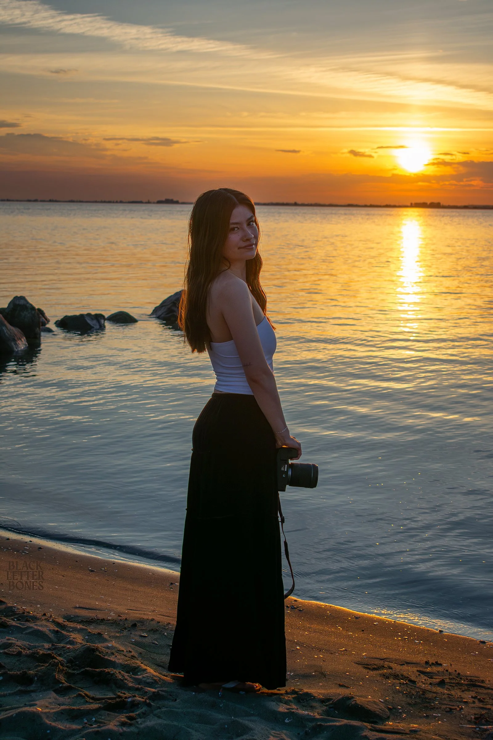 A woman standing on a beach holding a camera at sunset, with the water and rocks in the background.