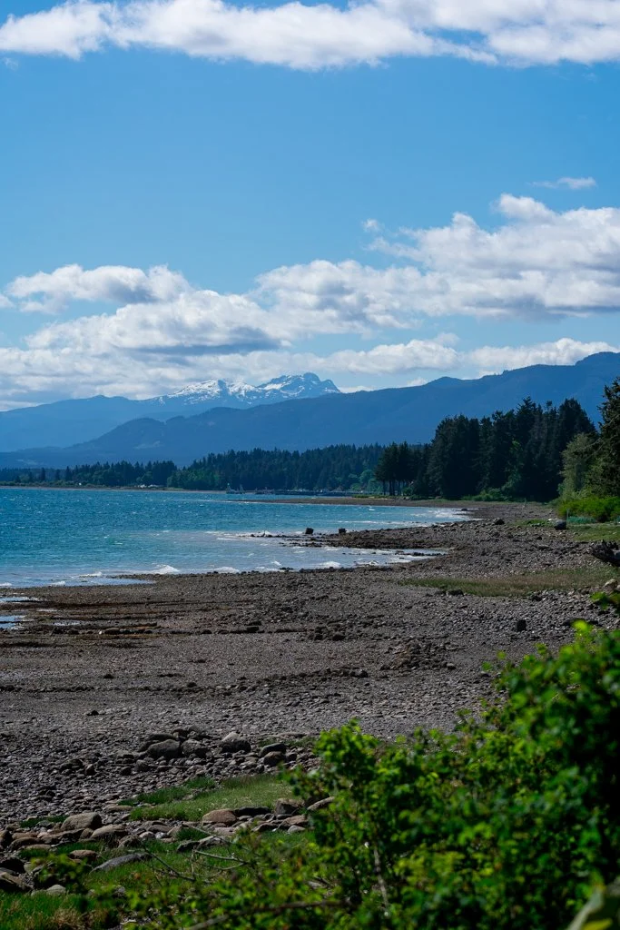 A rocky beach with a calm lake, green trees, and snow-capped mountains under a partly cloudy sky.