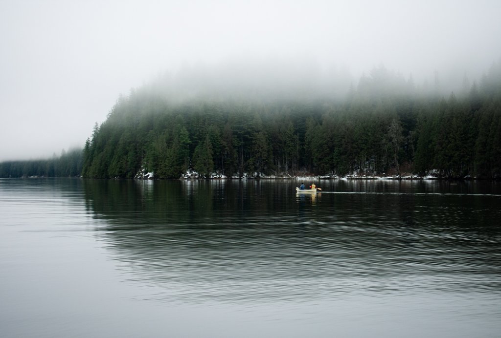 A small boat with two people paddling on a calm river surrounded by dense evergreen trees and fog-covered mountains in the background.