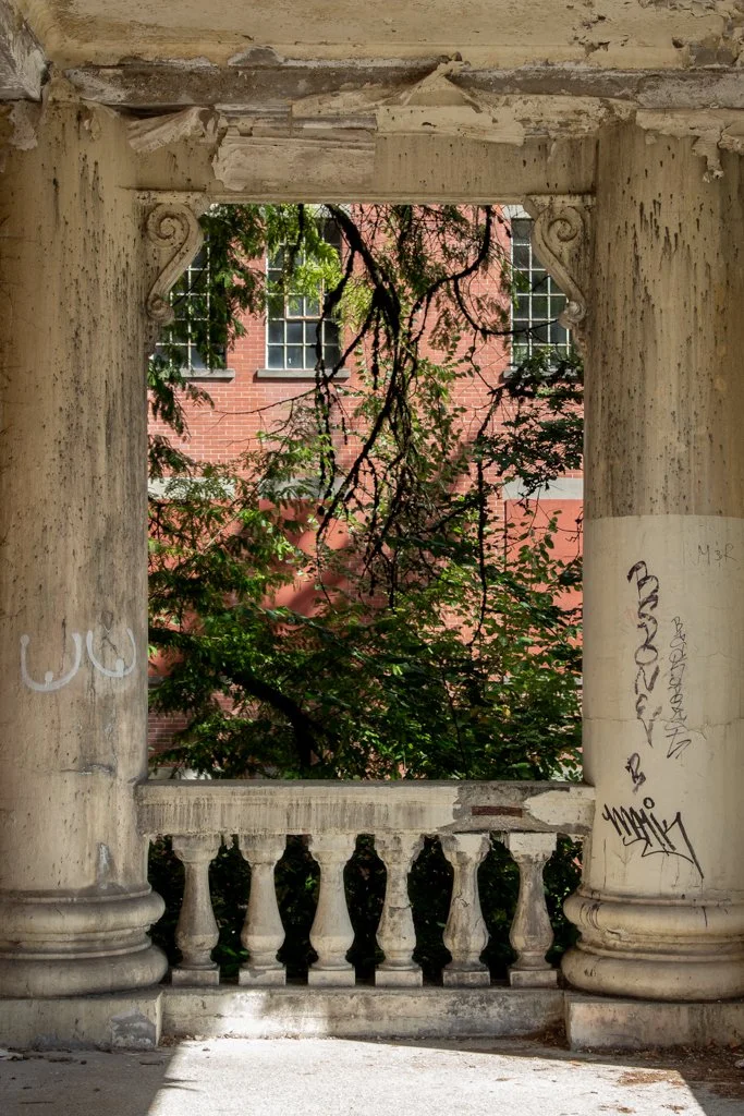 Old, weathered stone porch with a balustrade, graffiti on the columns, and lush trees and a brick building visible through an open window frame.