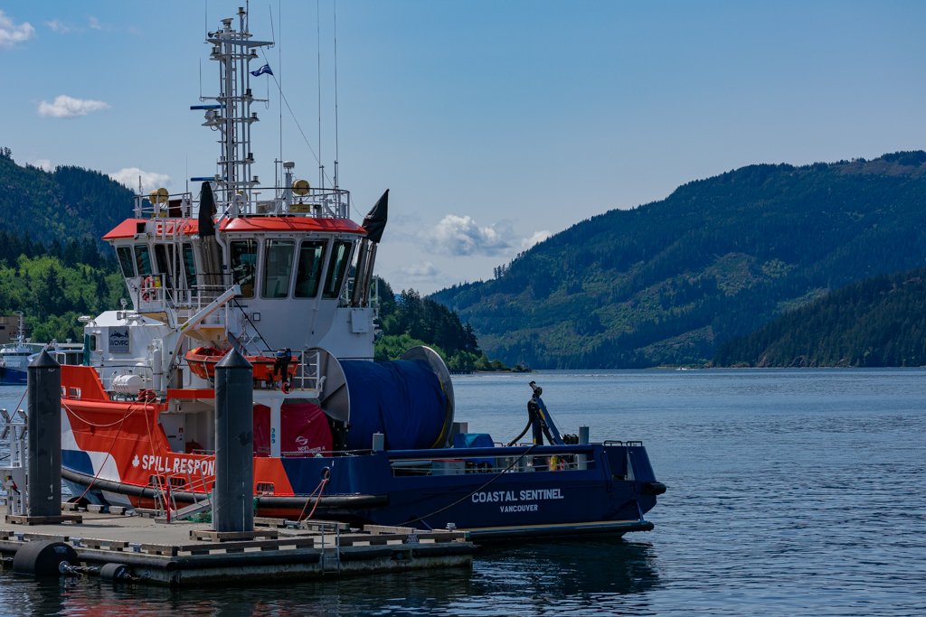 A coast guard rescue boat docked at a pier on a calm body of water, with forested hills in the background and a partly cloudy blue sky.