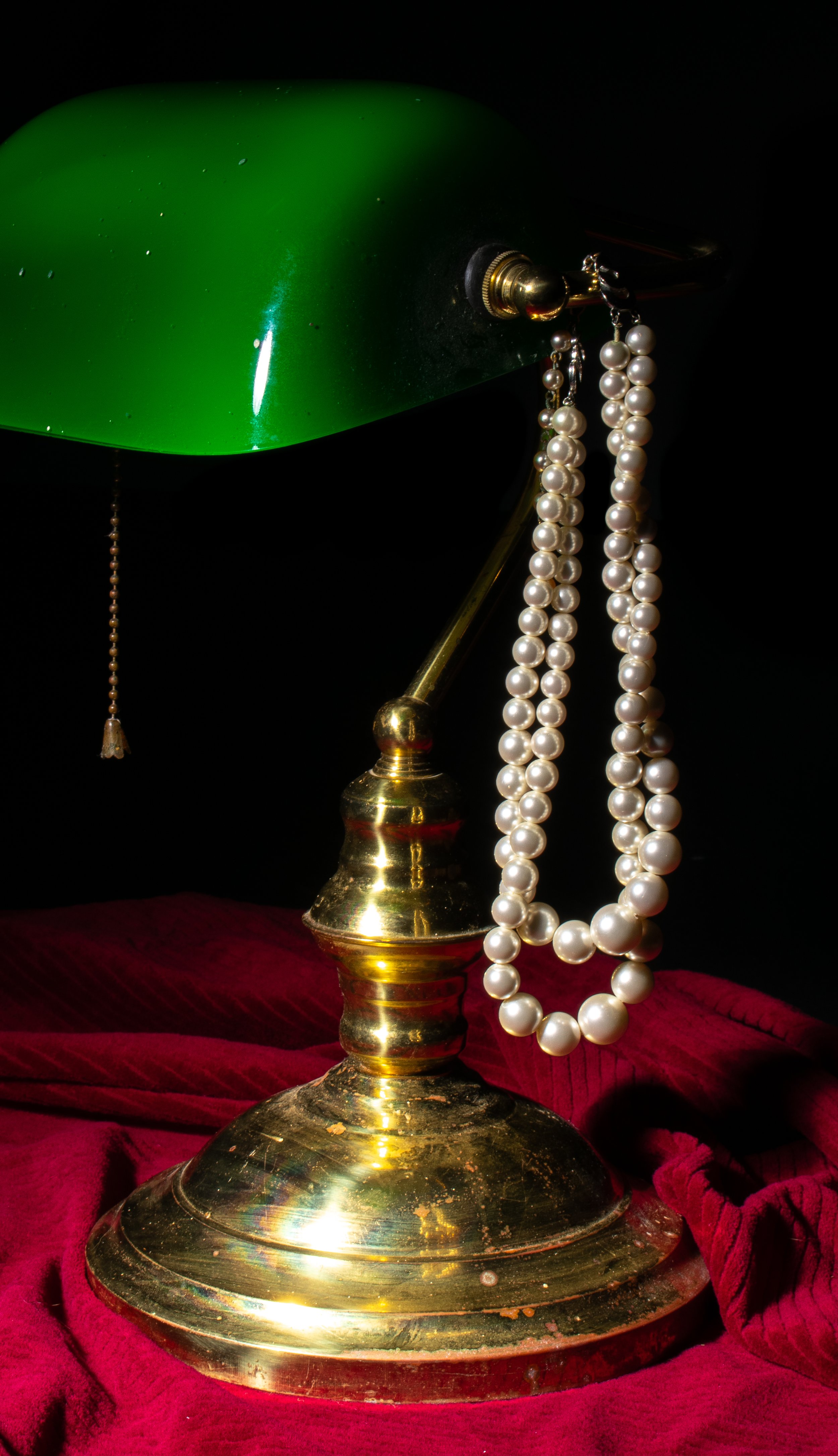 A vintage brass desk lamp with a green shade, a pearl necklace hanging from the lamp's fixture, and a red velvet cloth underneath.