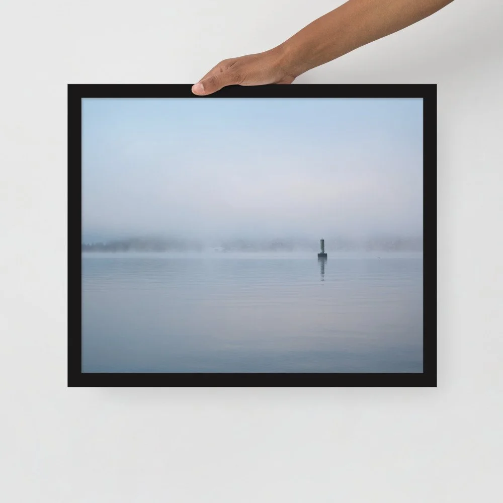 Hand holding a framed picture of a foggy body of water with a lighthouse in the distance.