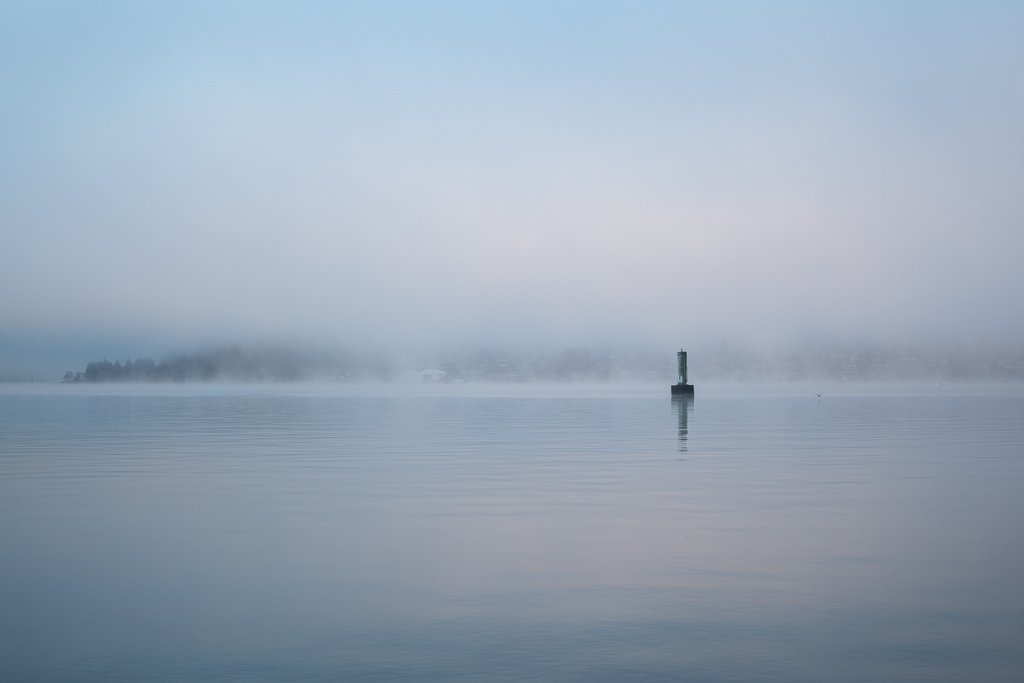 Calm body of water with a lighthouse in the distance, foggy atmosphere, and a faint shoreline with trees.