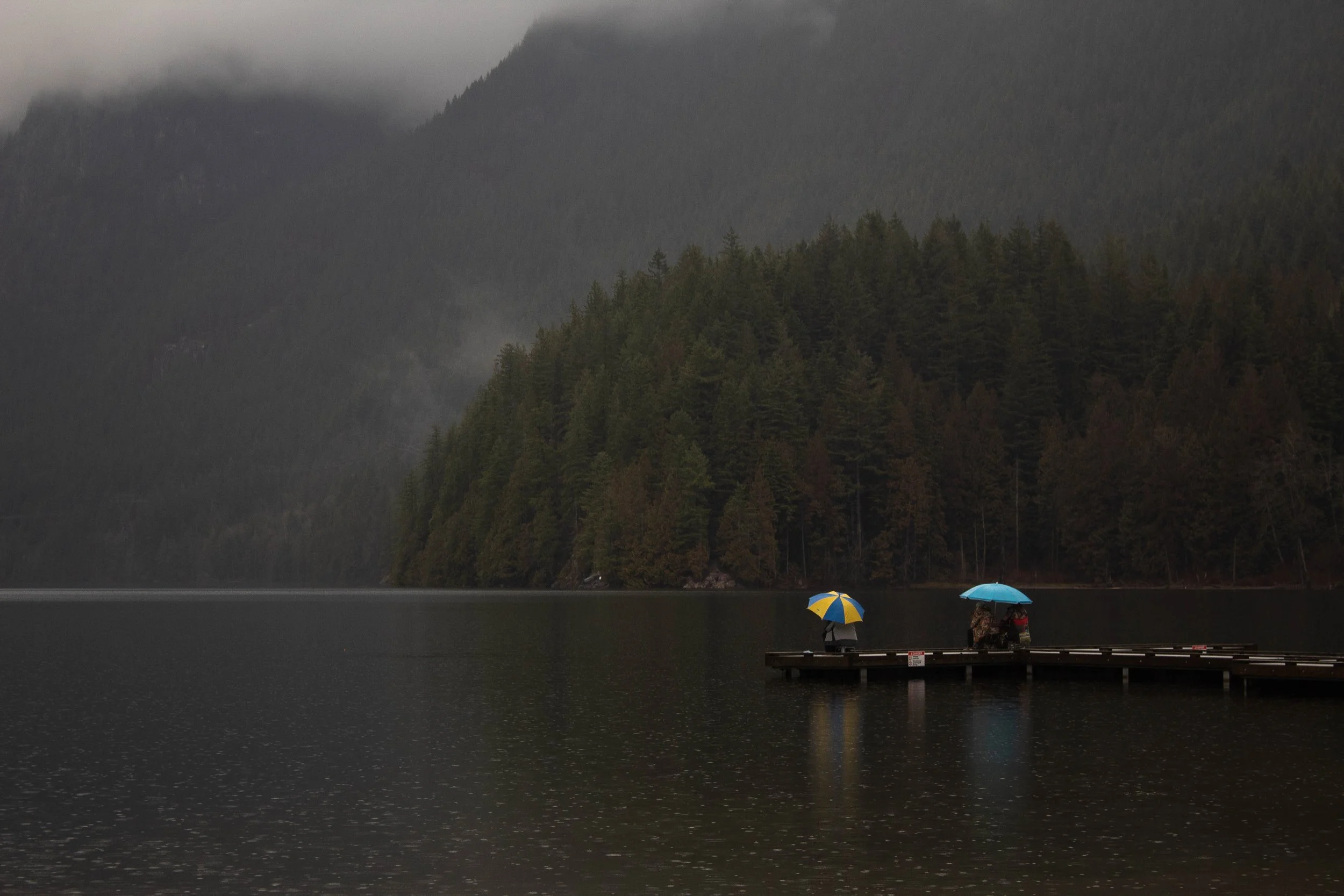A rainy day at a lake with two people on a dock, each holding an umbrella, surrounded by forested mountains.
