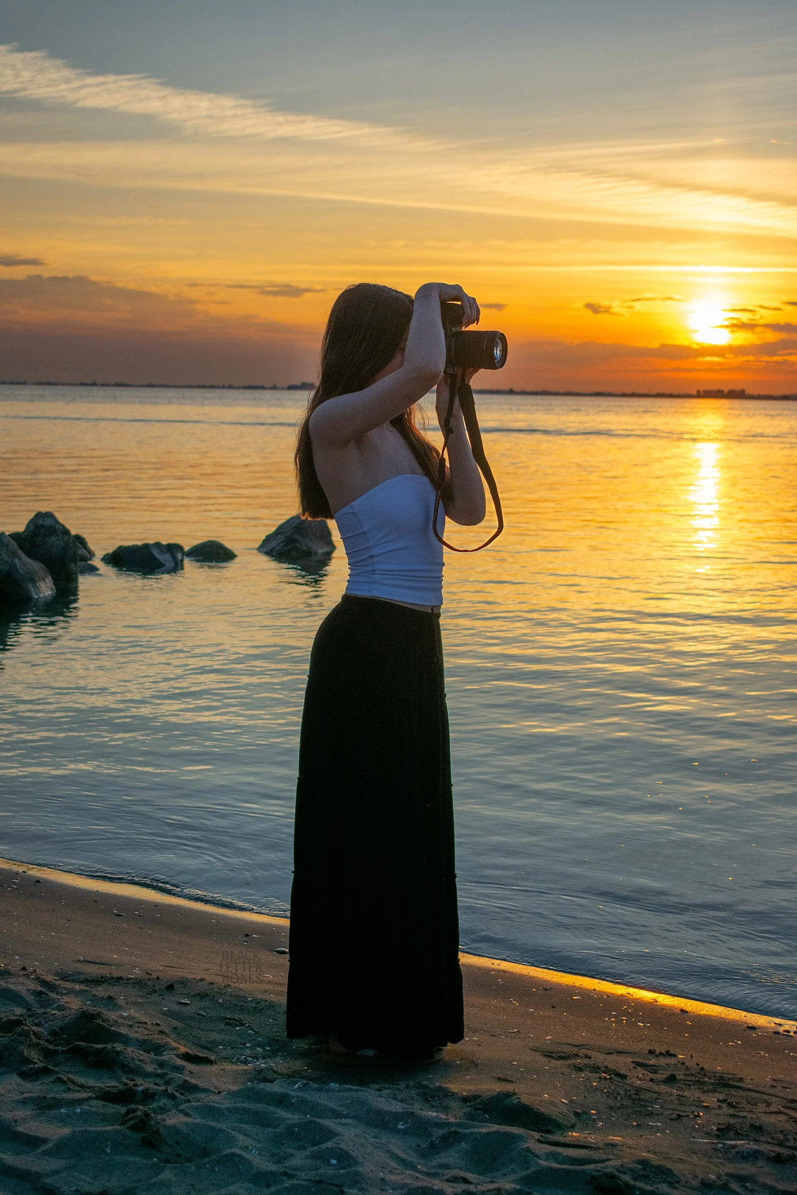 A woman standing on a beach taking a photo with her camera during sunset, with water and rocks in the background.