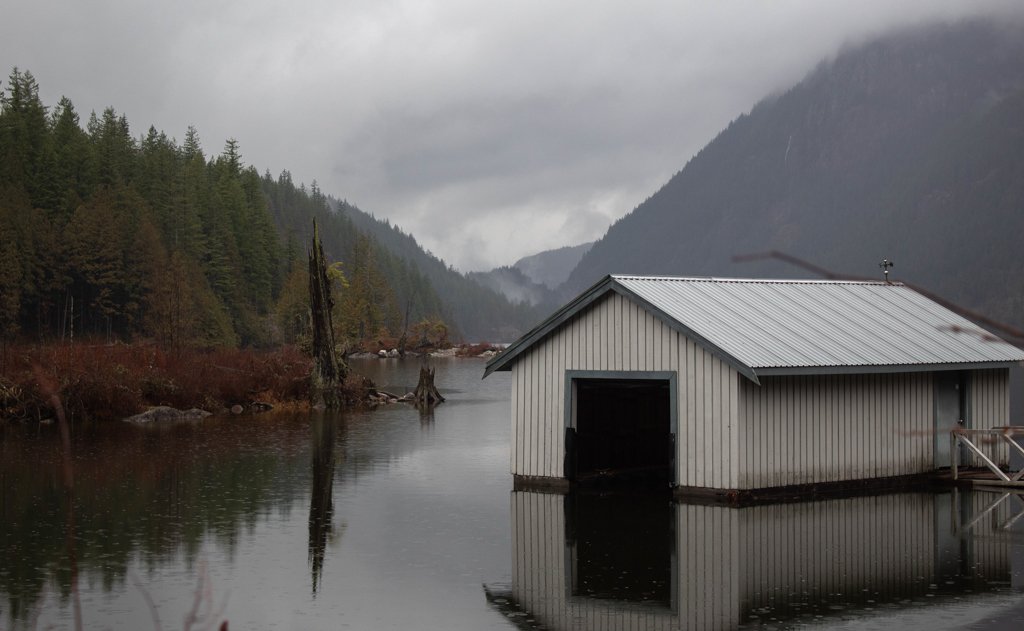 A gray metal boathouse reflects on calm water, surrounded by forested mountains under a cloudy sky.